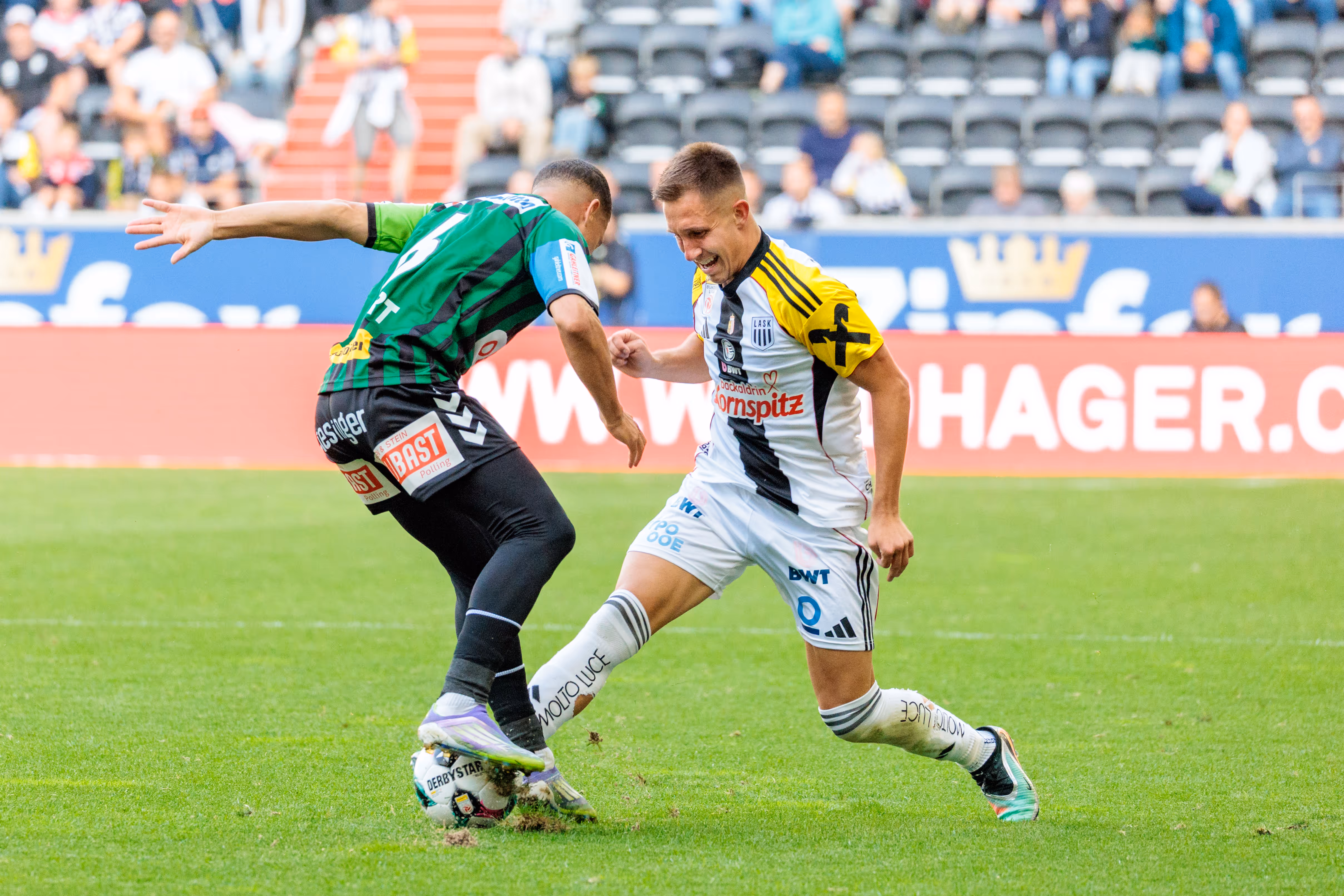 Zwei Fußballspieler, einer im weiß-schwarzen LASK-Trikot und einer im grün-schwarzen SV Ried-Trikot, liefern sich ein hartes Duell um den Ball in einem Bundesligaspiel
