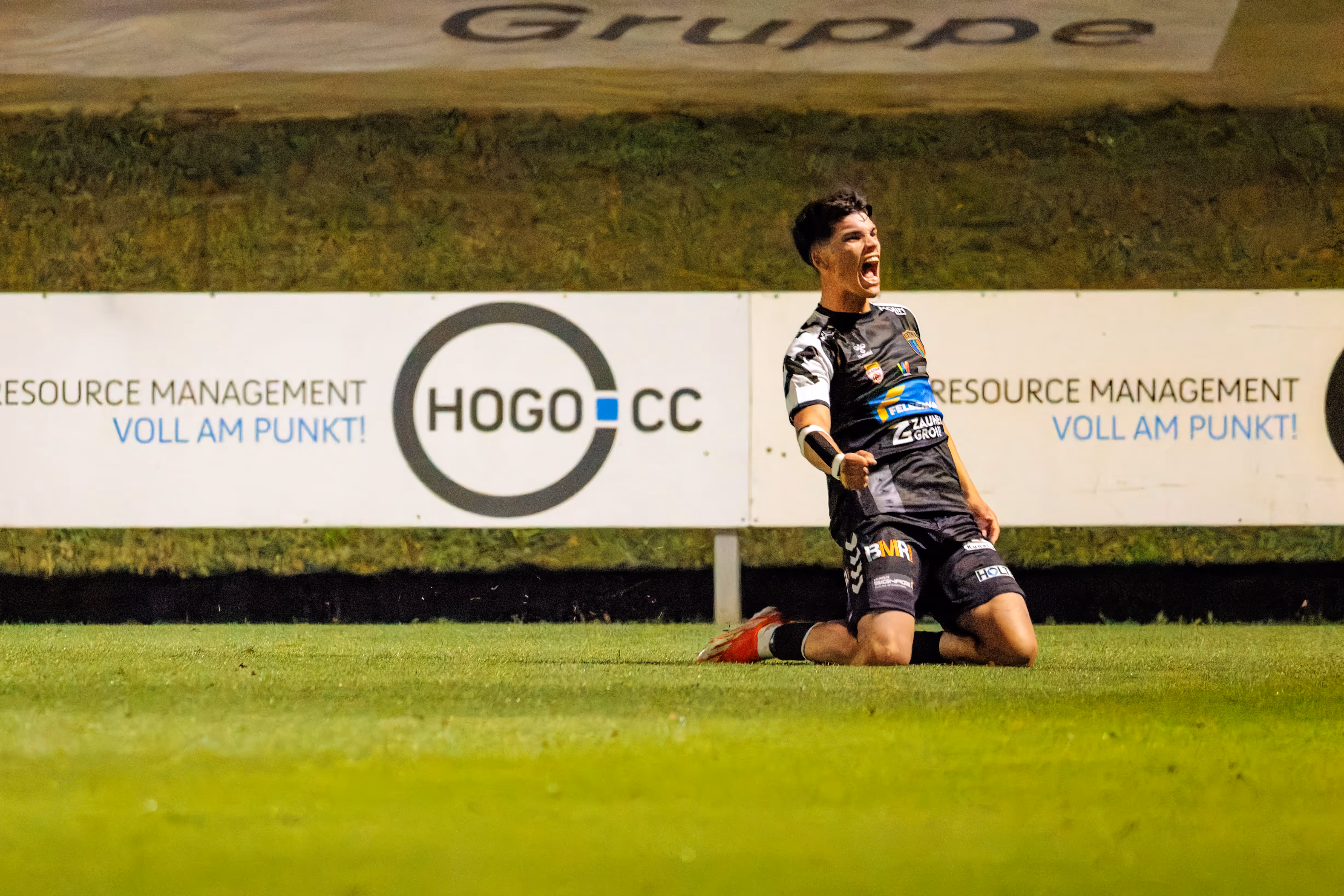 Ein Spieler der SPG HOGO Wels jubelt kniend und mit geballten Fäusten vor einer Werbebande mit dem „HOGO CC“-Logo über einen wichtigen Ausgleichstreffer