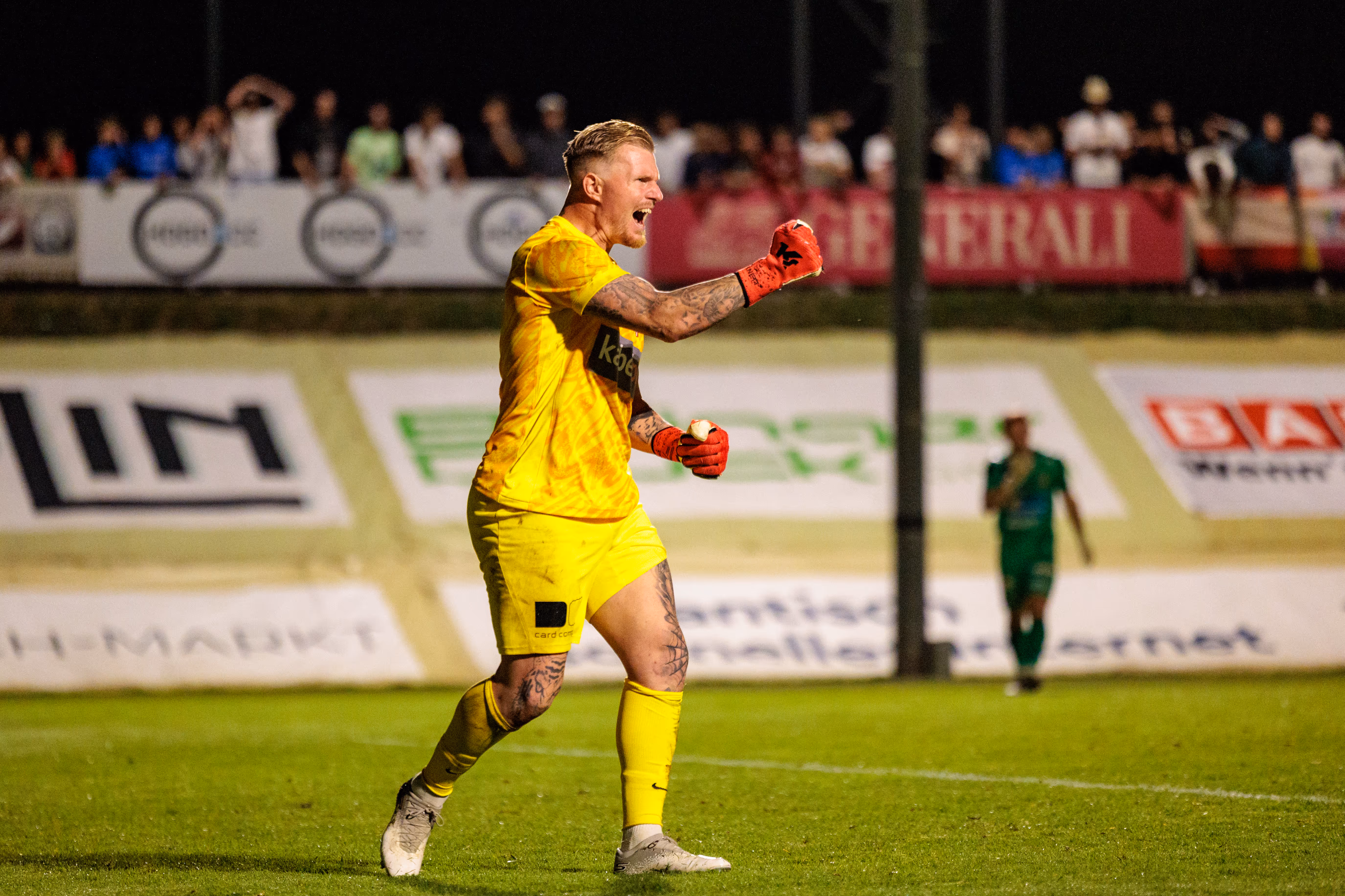 Ein leidenschaftlich jubelnder Fußballtorwart in leuchtend gelbem Trikot und roten Handschuhen feiert mit geöffnetem Mund und geballten Fäusten auf dem Spielfeld nach einem Elfmeterschießen