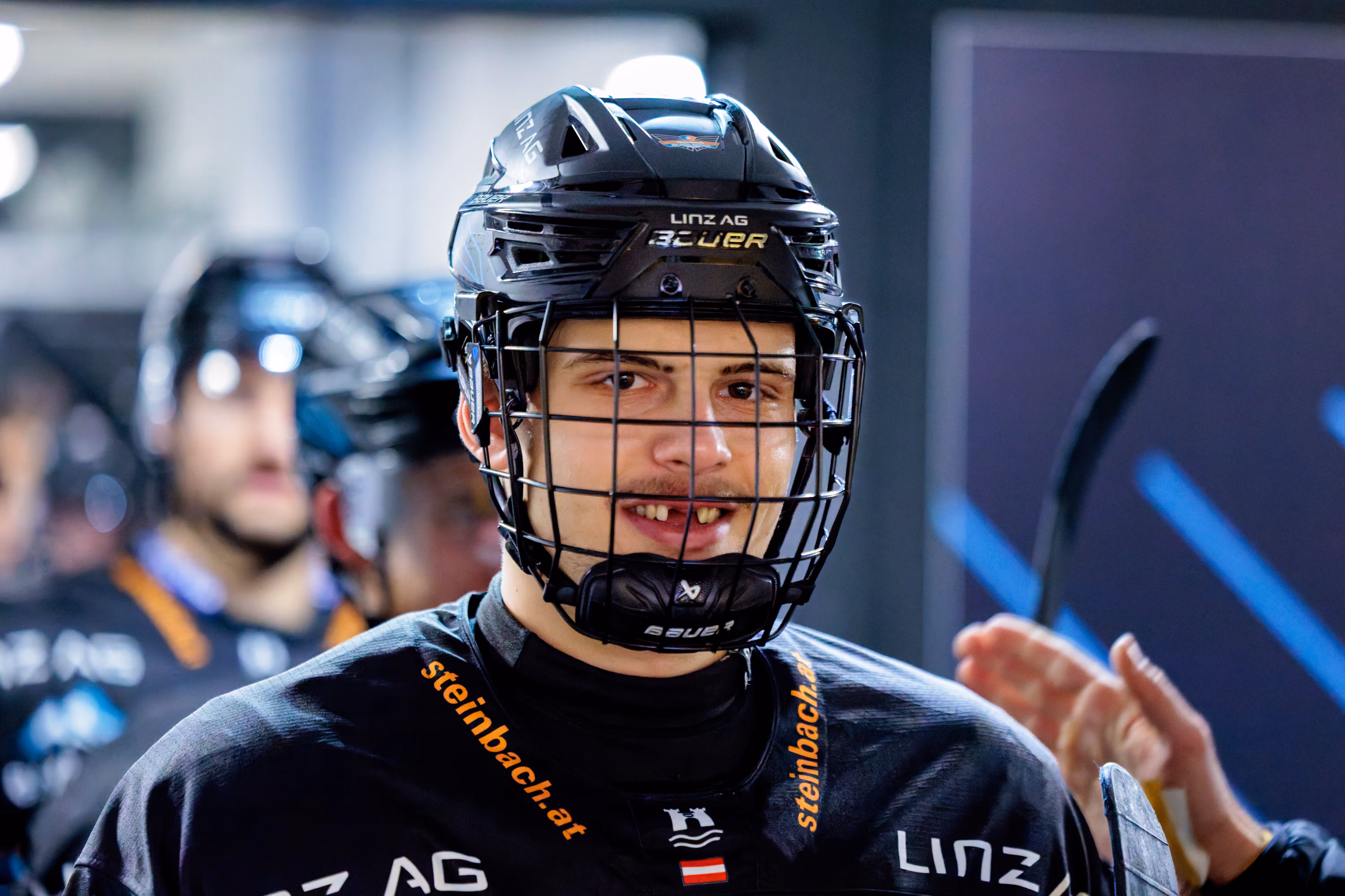 Porträt eines jungen Eishockeyspielers der Steinbach Black Wings Linz, der mit seinem schwarzen Helm und Gitter leicht in die Kamera lächelt