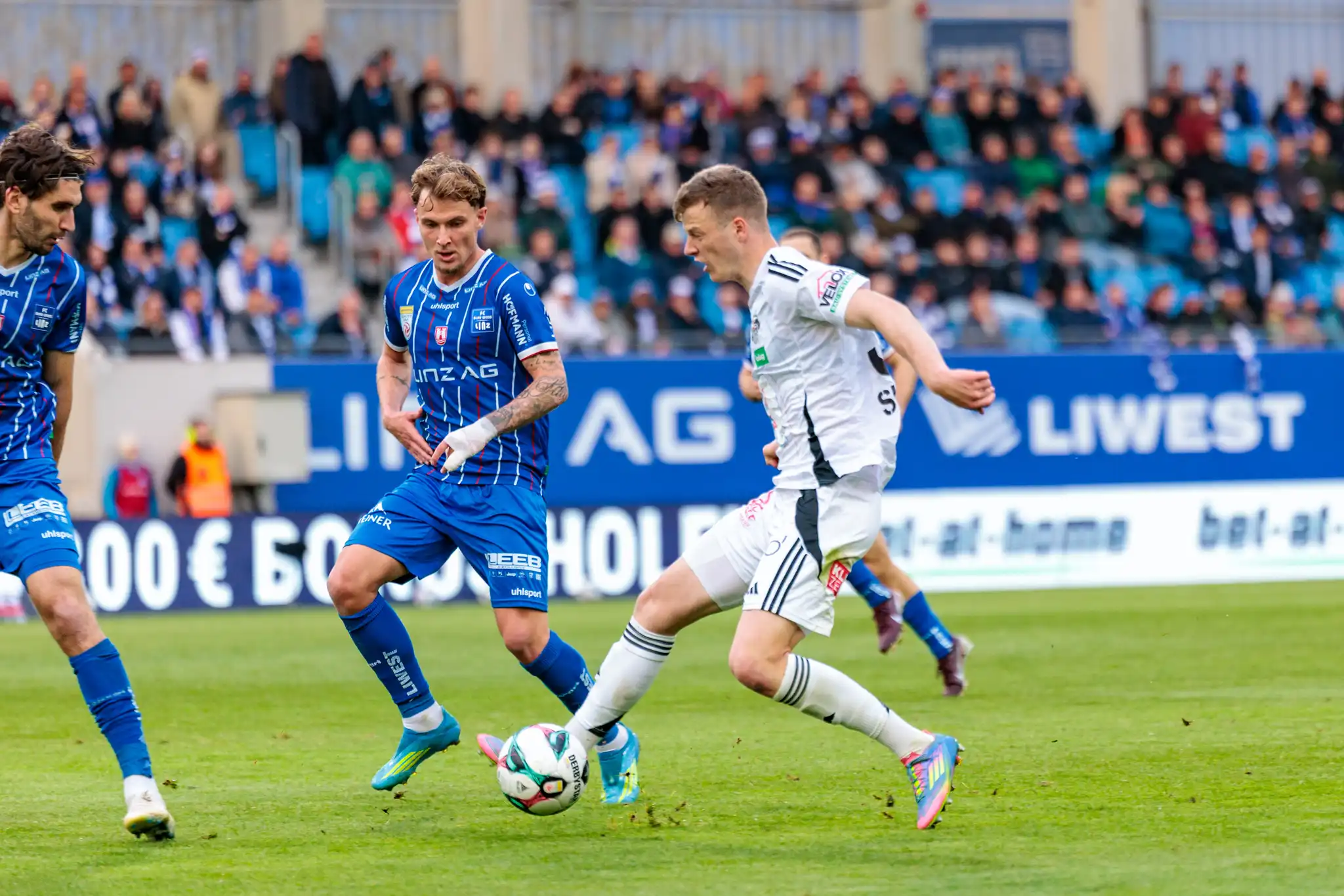 Ein Spieler des Wolfsberger AC kämpft in einem intensiven Zweikampf um den Ball gegen zwei Gegenspieler des FC Blau-Weiß Linz