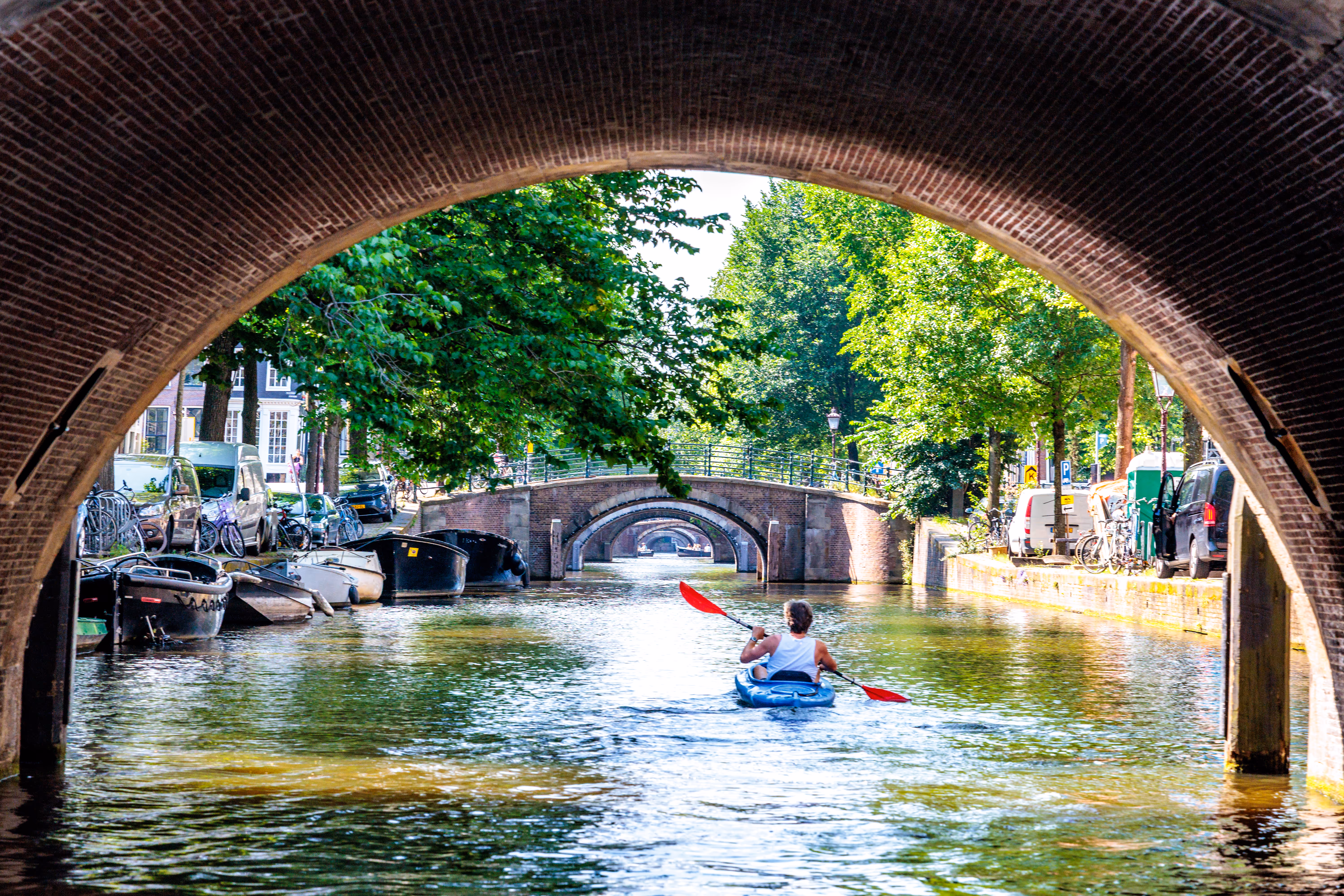 Eine Person in einem blauen Kajak paddelt unter einer historischen Backsteinbrücke auf einem sonnigen Kanal in Amsterdam, umgeben von grünen Bäumen und urbaner Architektur 