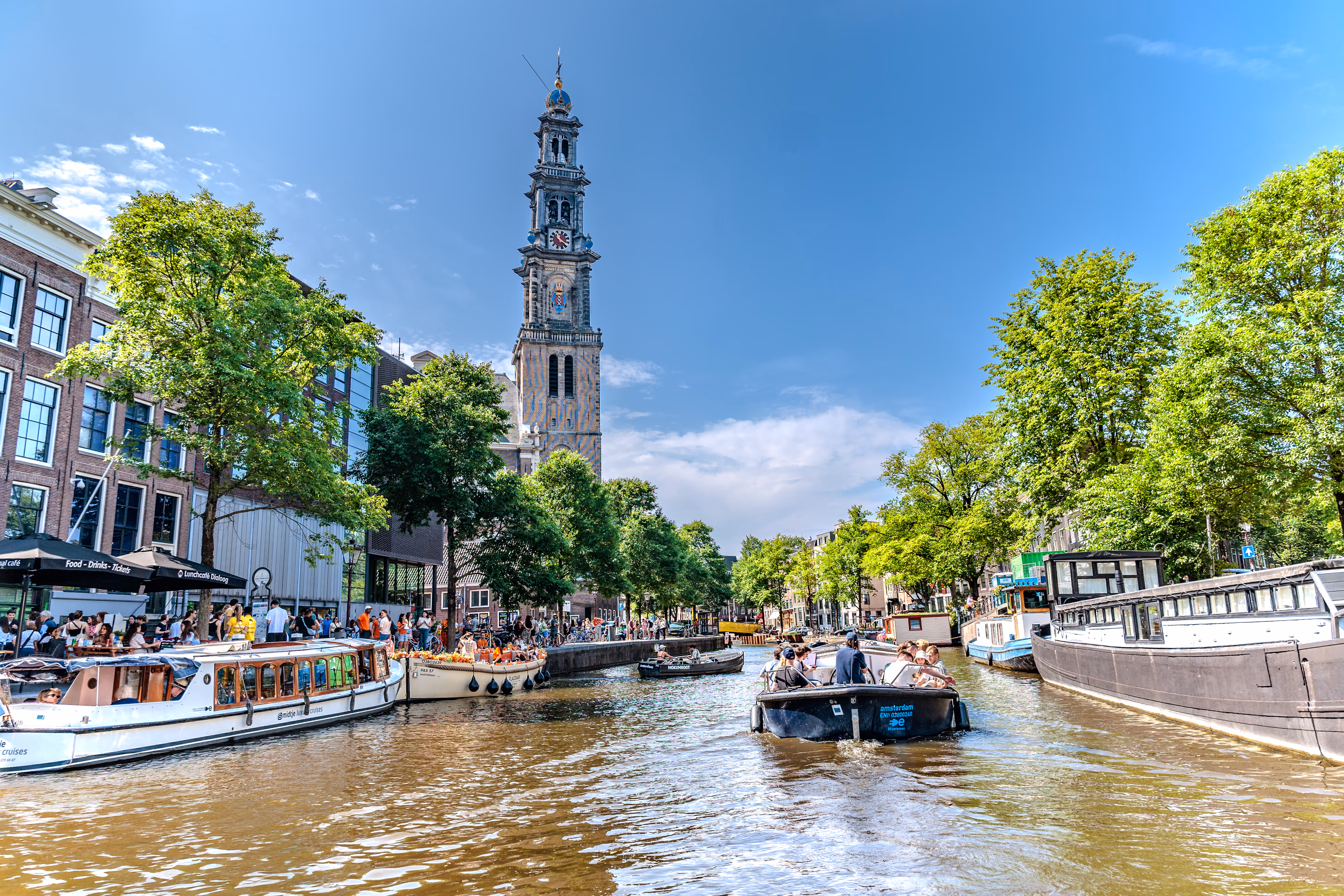 Blick auf einen belebten Amsterdamer Kanal mit vielen Booten und der historischen Westerkerk unter strahlend blauem Himmel 