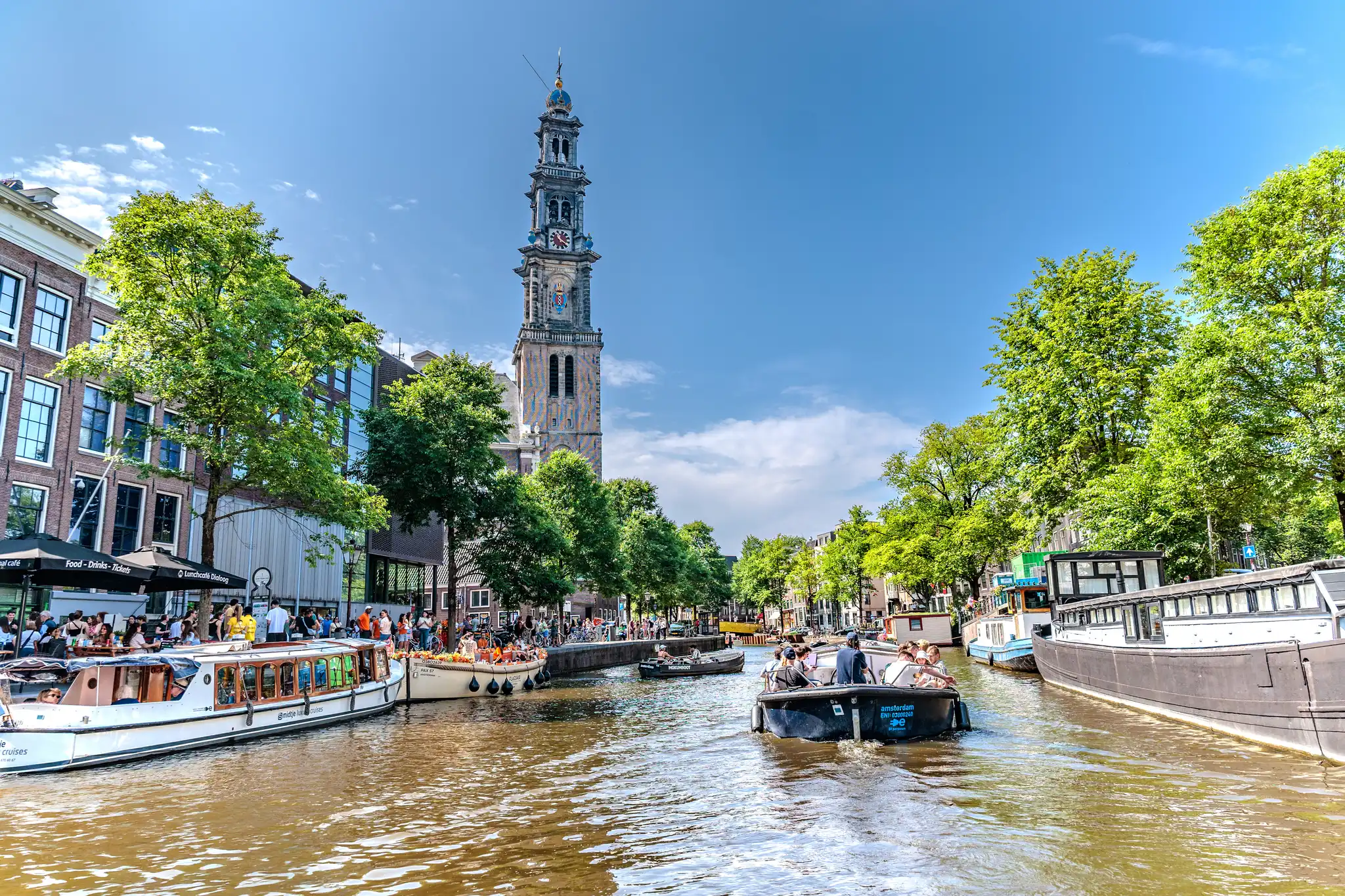 Blick auf einen belebten Amsterdamer Kanal mit vielen Booten und der historischen Westerkerk unter strahlend blauem Himmel