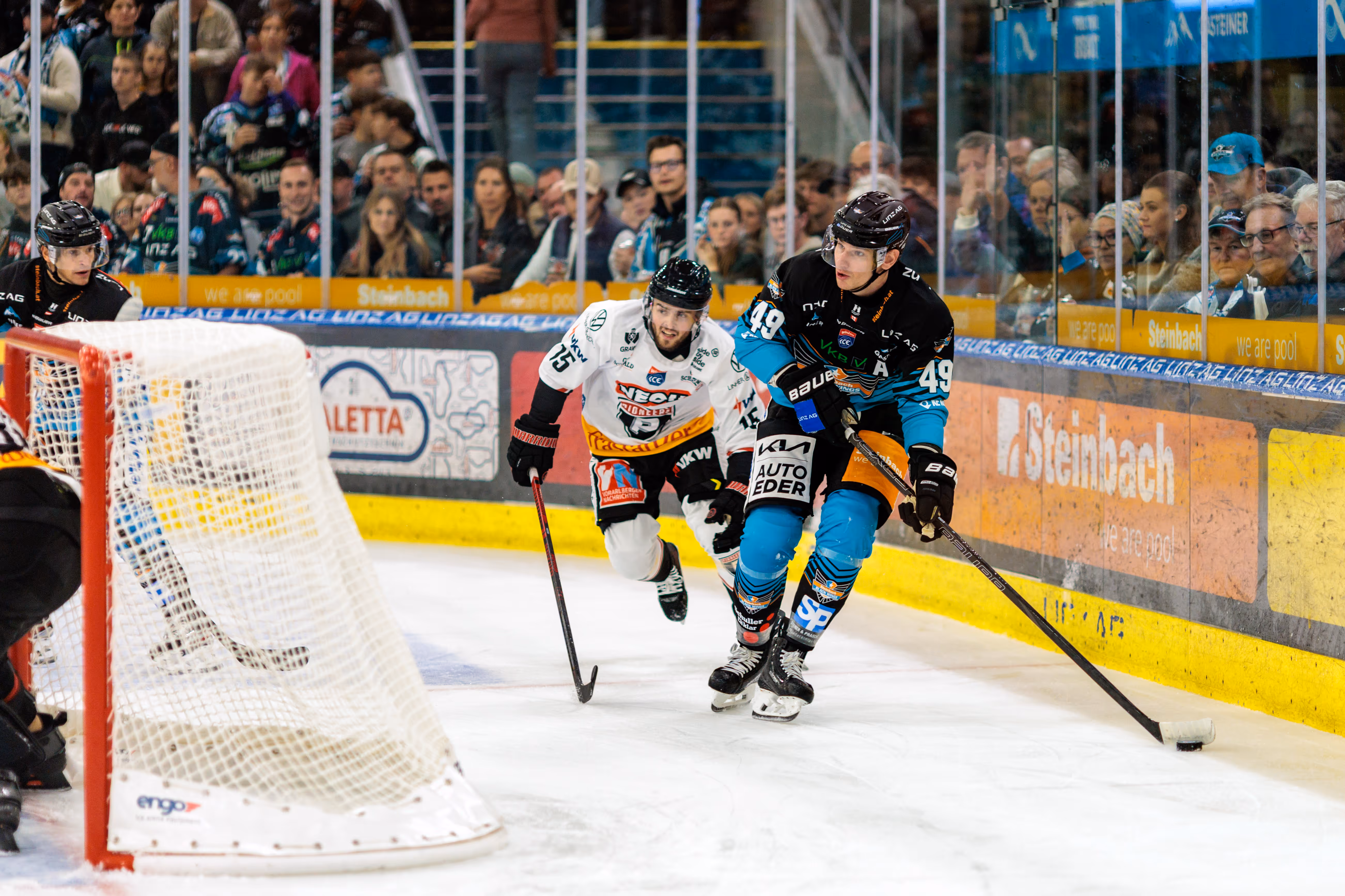 Eishockeyspieler Graham Knott von den Steinbach Black Wings Linz im teal-schwarzen Trikot mit der Nummer 49 führt den Puck über das Eis, während Mark Mussbacher der Pioneers Vorarlberg mit der Nummer 15 ihm folgt