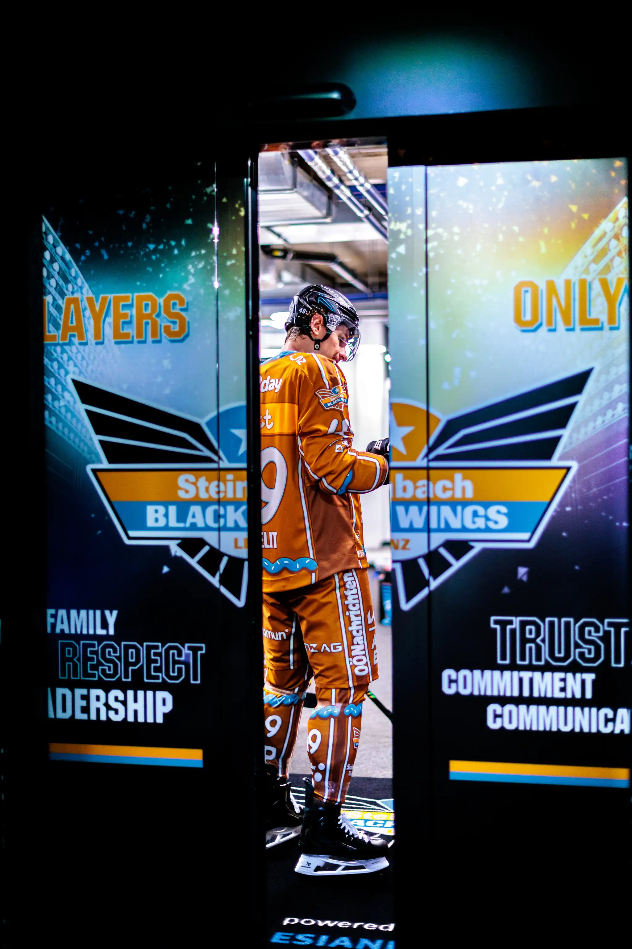 Eishockeyspieler Graham Knott der Steinbach Black Wings Linz in orangem Trikot mit der Nummer 49 steht nachdenklich in einem beleuchteten Spielertunnel mit Team-Logos