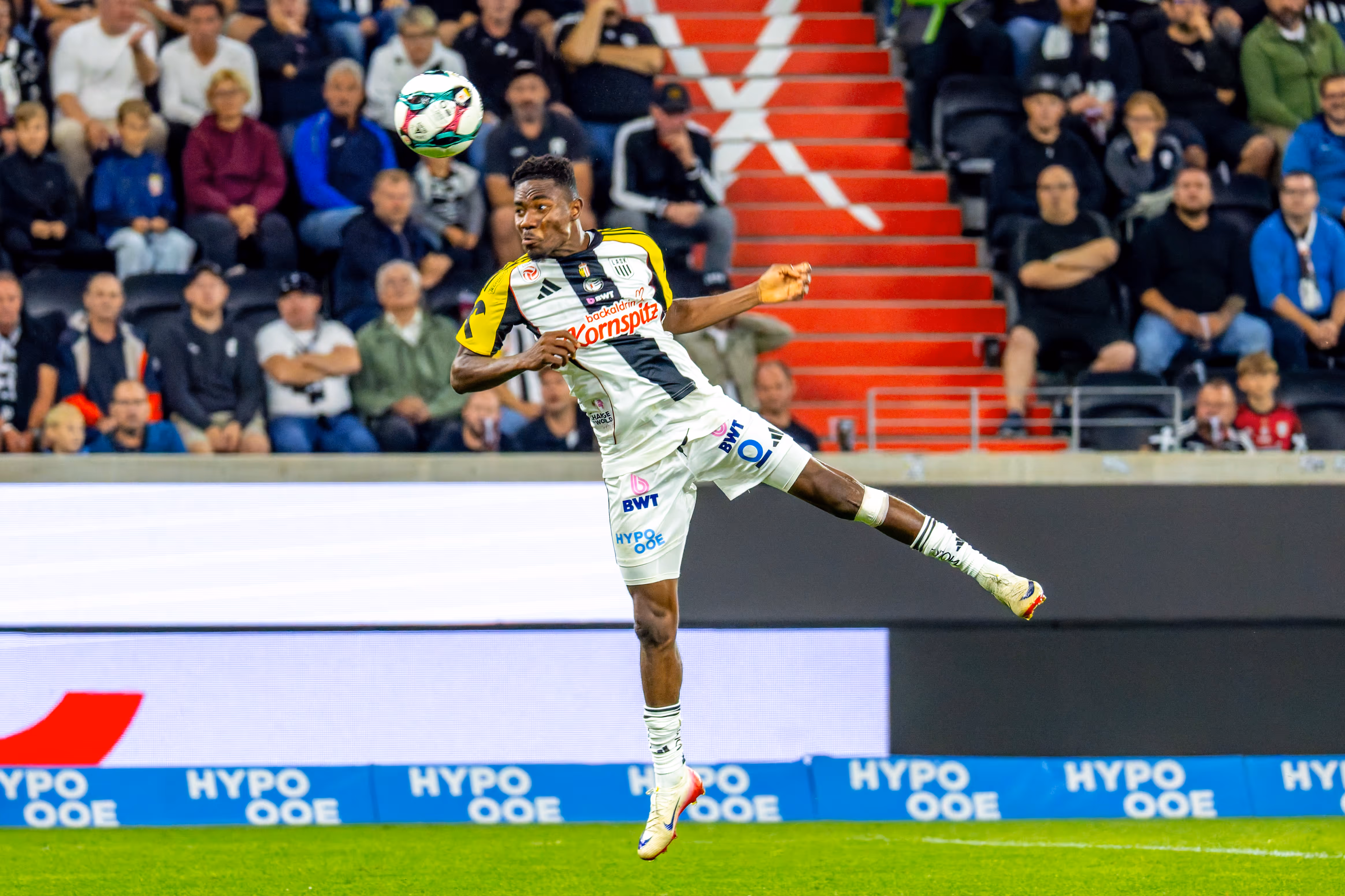 Ein LASK-Spieler im weiß-gelben Trikot springt im Stadion zum Kopfball, während Fans auf den Rängen zu sehen sind