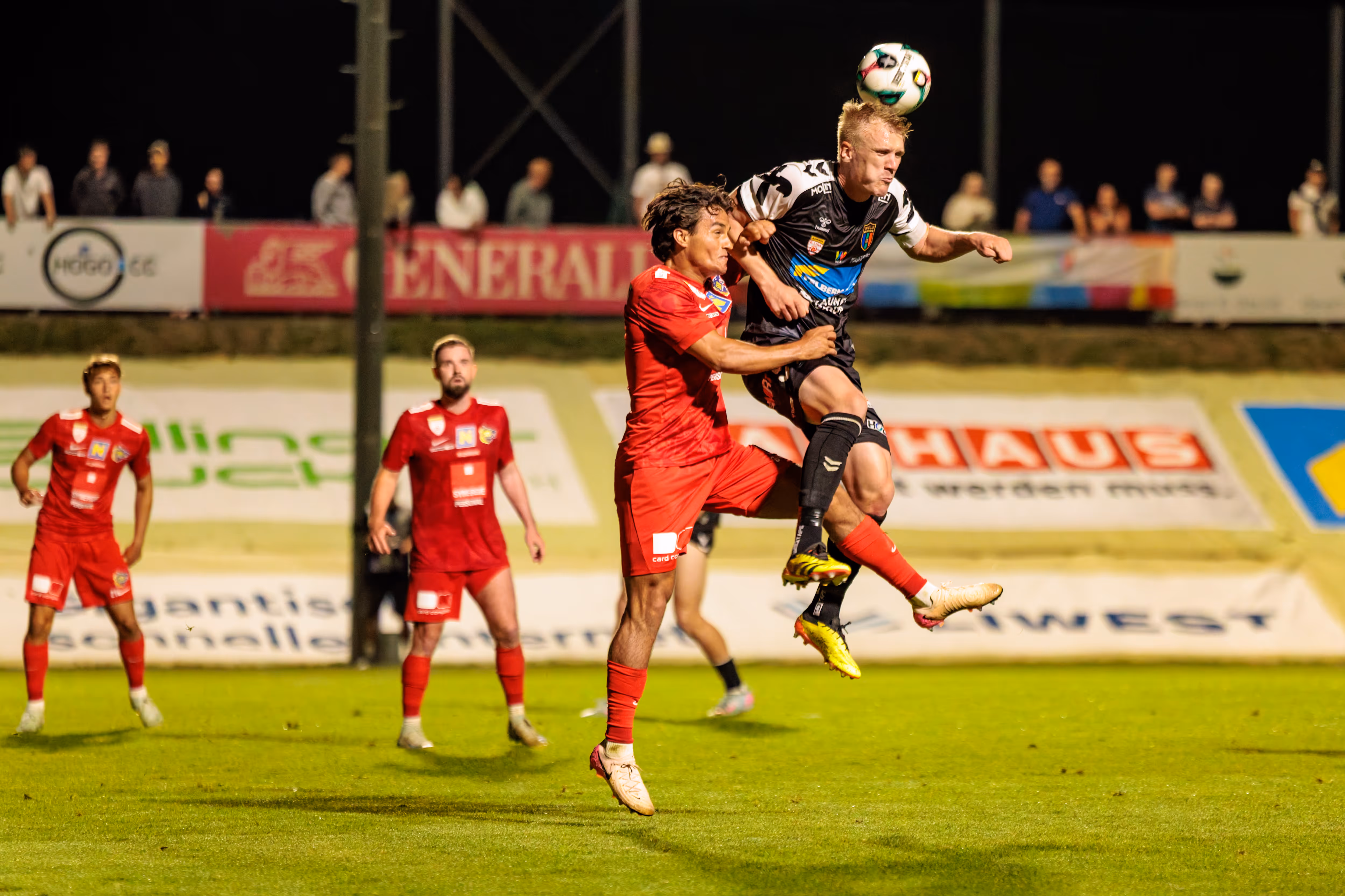 Ein intensiver Kopfball-Zweikampf zwischen zwei Fußballspielern in der Luft während eines abendlichen Matches, bei dem der Spieler von Herta Wels den Ball köpft
