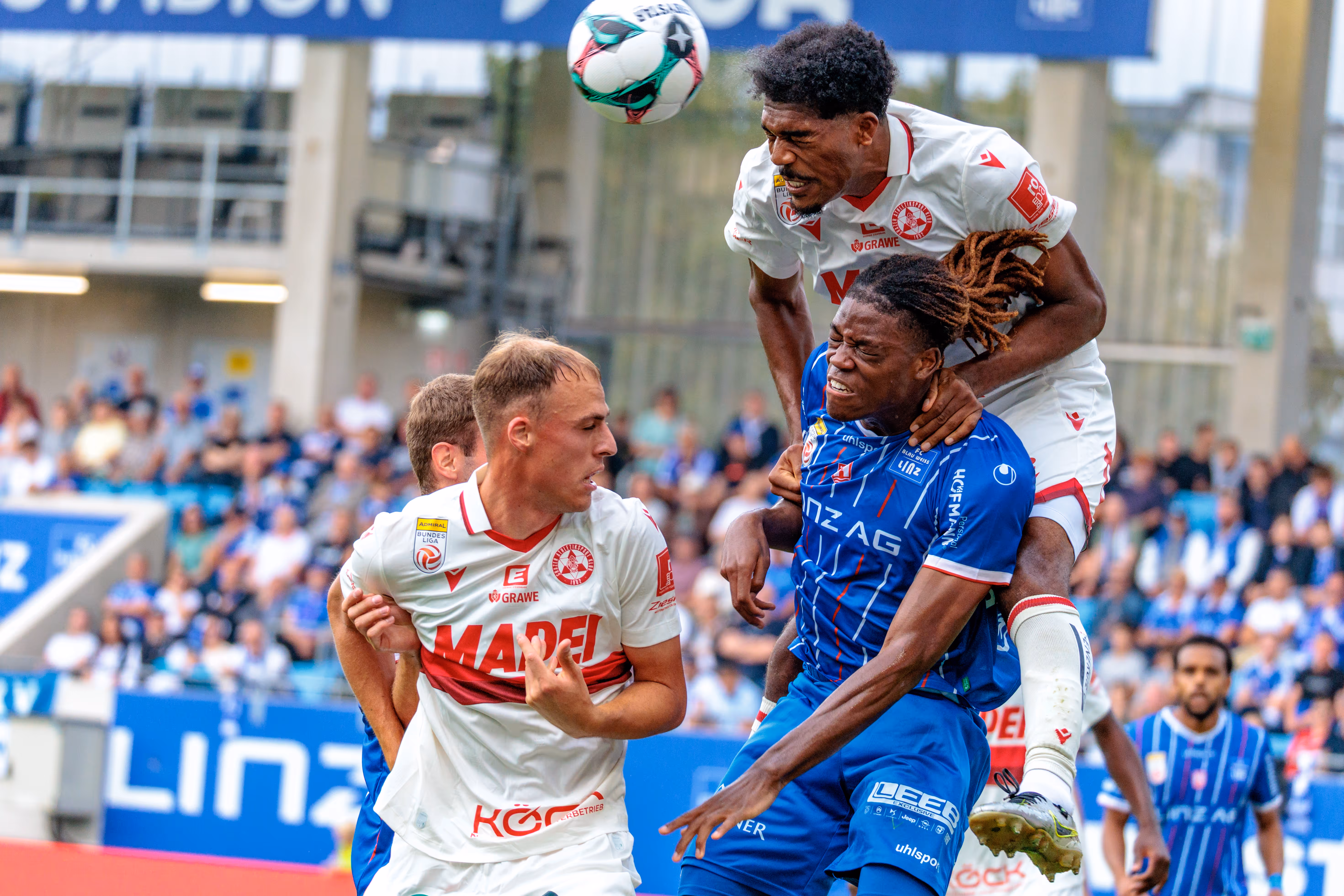 Der GAK-Spieler Donovan Pines steigt in einem dynamischen Kopfballduell über den Blau-Weiß Linz-Spieler Alem Pasic, während ein weiterer GAK-Akteur die Szene im Vordergrund beobachtet