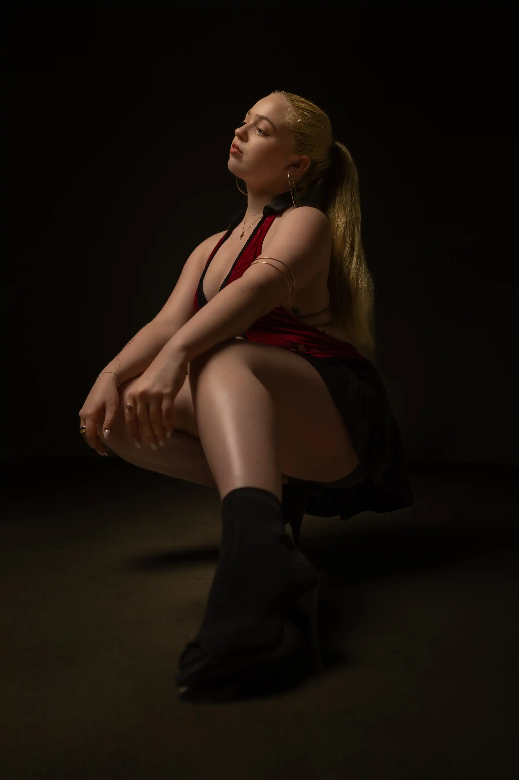 Eine blonde Frau in rotem Oberteil und dunklem Rock kniet mit aufwärts gerichtetem Blick, im Seitenlicht vor dunklem Hintergrund 