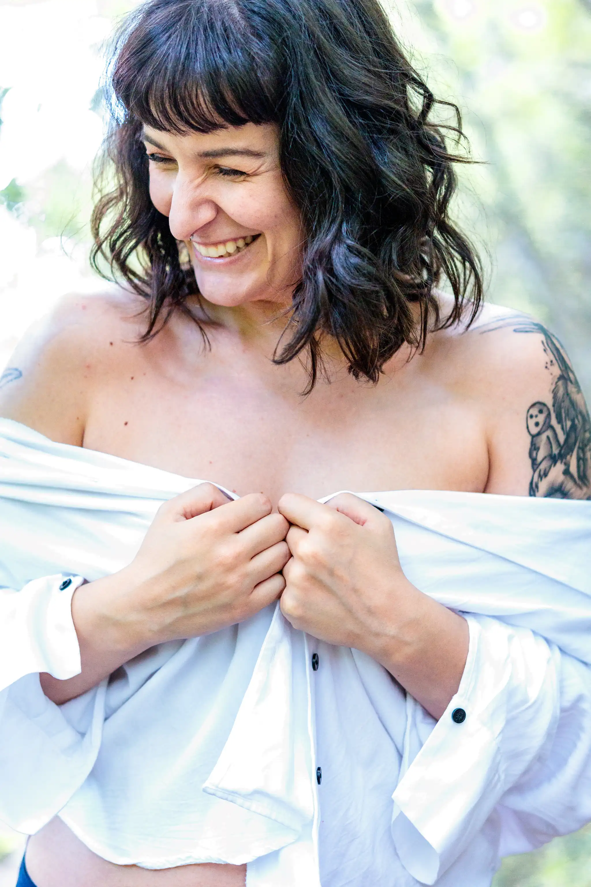 Eine lächelnde Frau mit dunklen Haaren und markanten Tattoos trägt ein schulterfreies, weißes Hemd vor einem weichen, grünen Hintergrund 