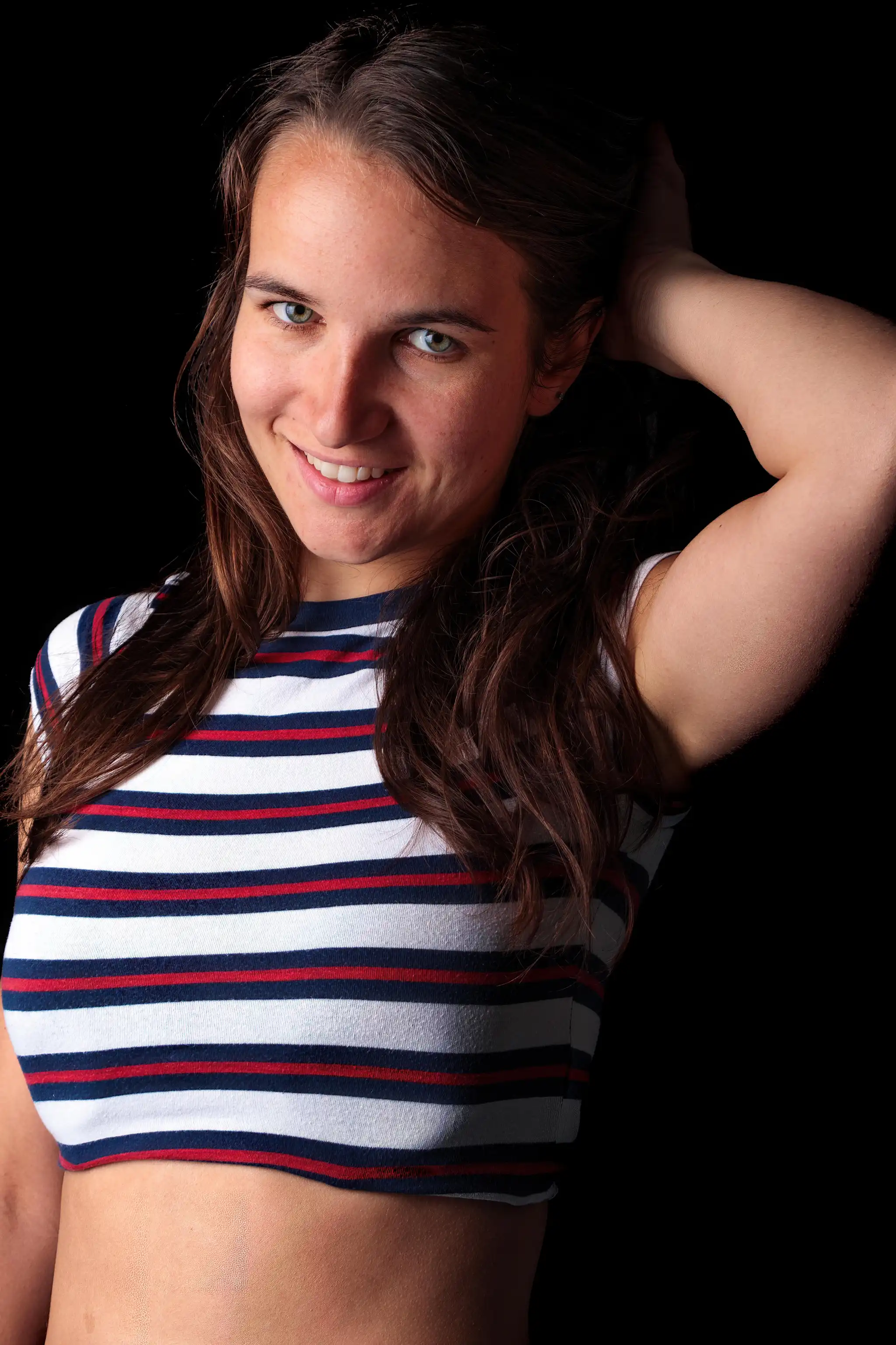 Eine lächelnde junge Frau mit braunen Haaren und grünen Augen trägt ein gestreiftes Crop-Top vor schwarzem Hintergrund 