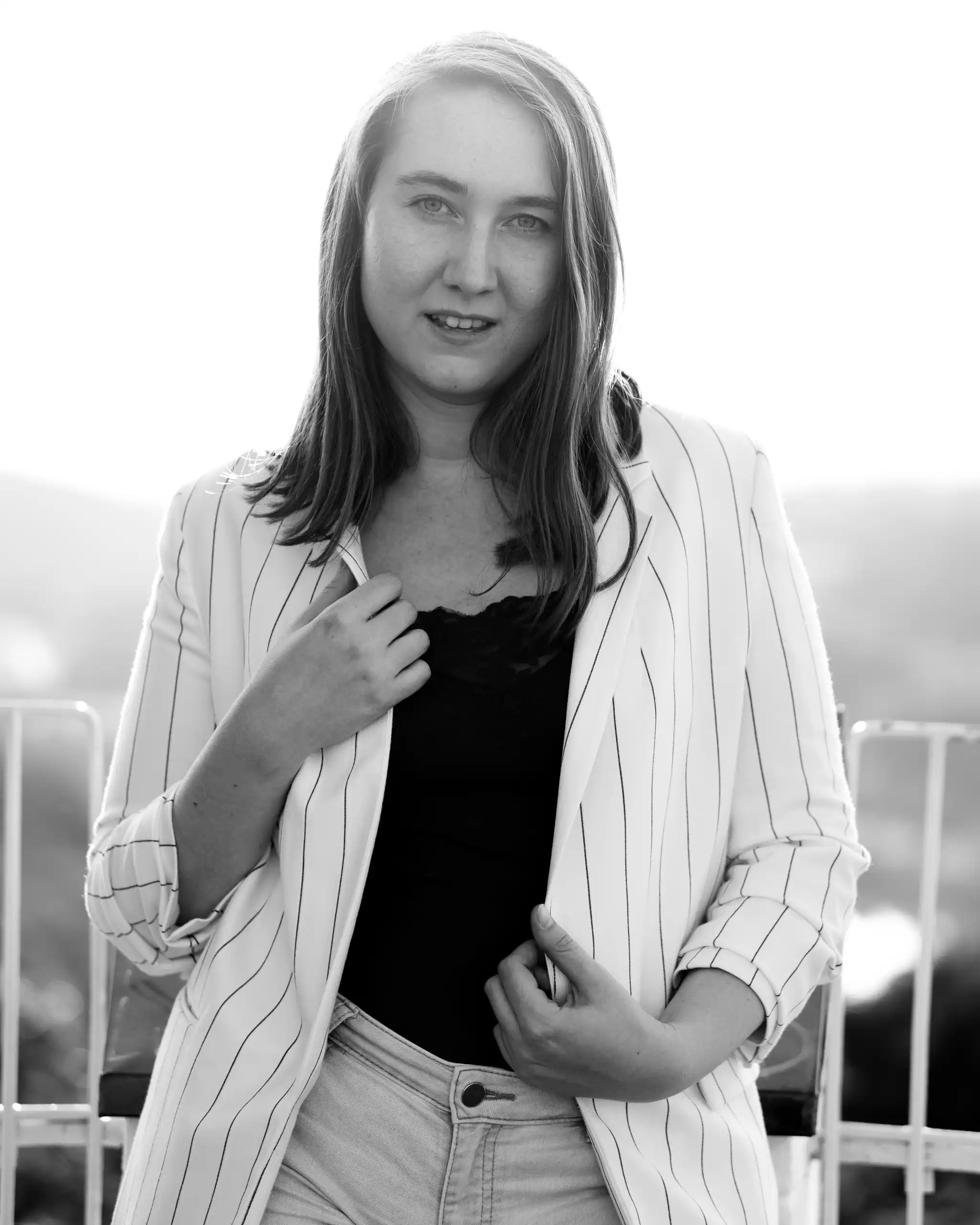 Porträt einer lächelnden jungen Frau in einem gestreiften Blazer und heller Jeans, aufgenommen im Gegenlicht im Freien 