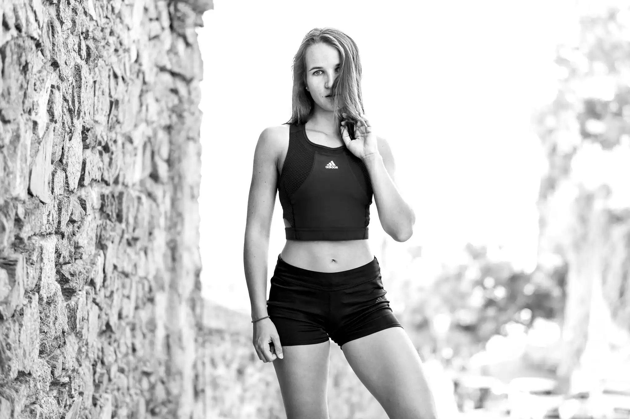 Schwarz-Weiß-Porträt von Anna Wirth in Adidas Sportkleidung, die vor einer rauen Steinmauer steht 