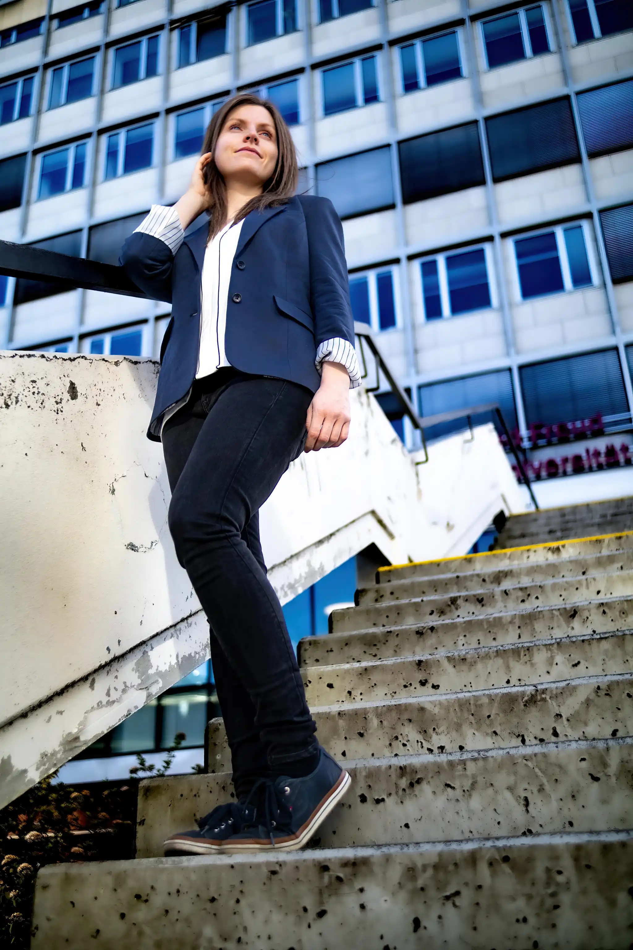 Eine junge Geschäftsfrau im blauen Blazer steht auf einer Betontreppe vor einem modernen Bürogebäude und blickt nachdenklich nach oben 
