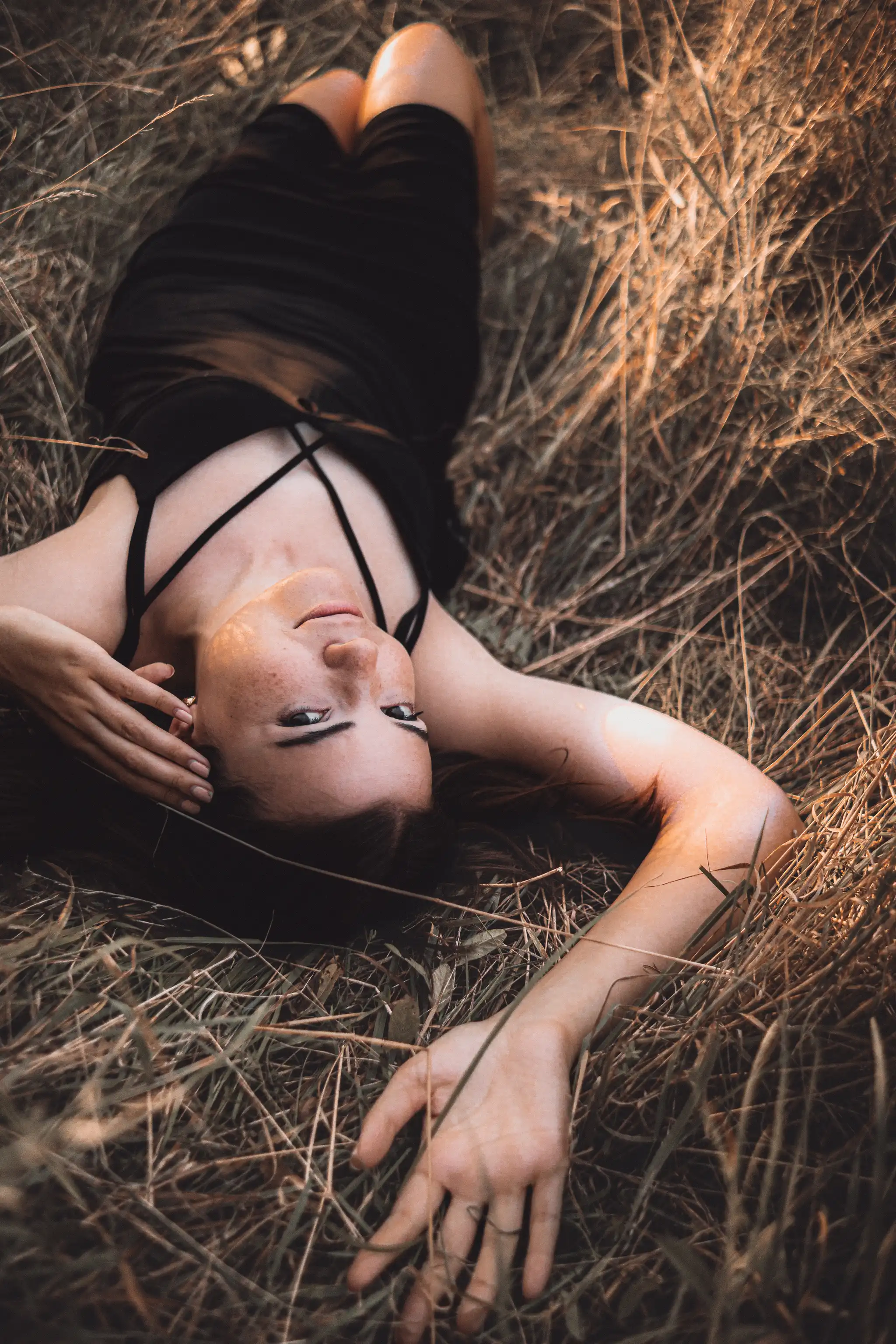 Eine junge Frau liegt in einem Feld aus trockenem Gras, ihr Gesicht von der Abendsonne beleuchtet, während sie in die Kamera blickt 