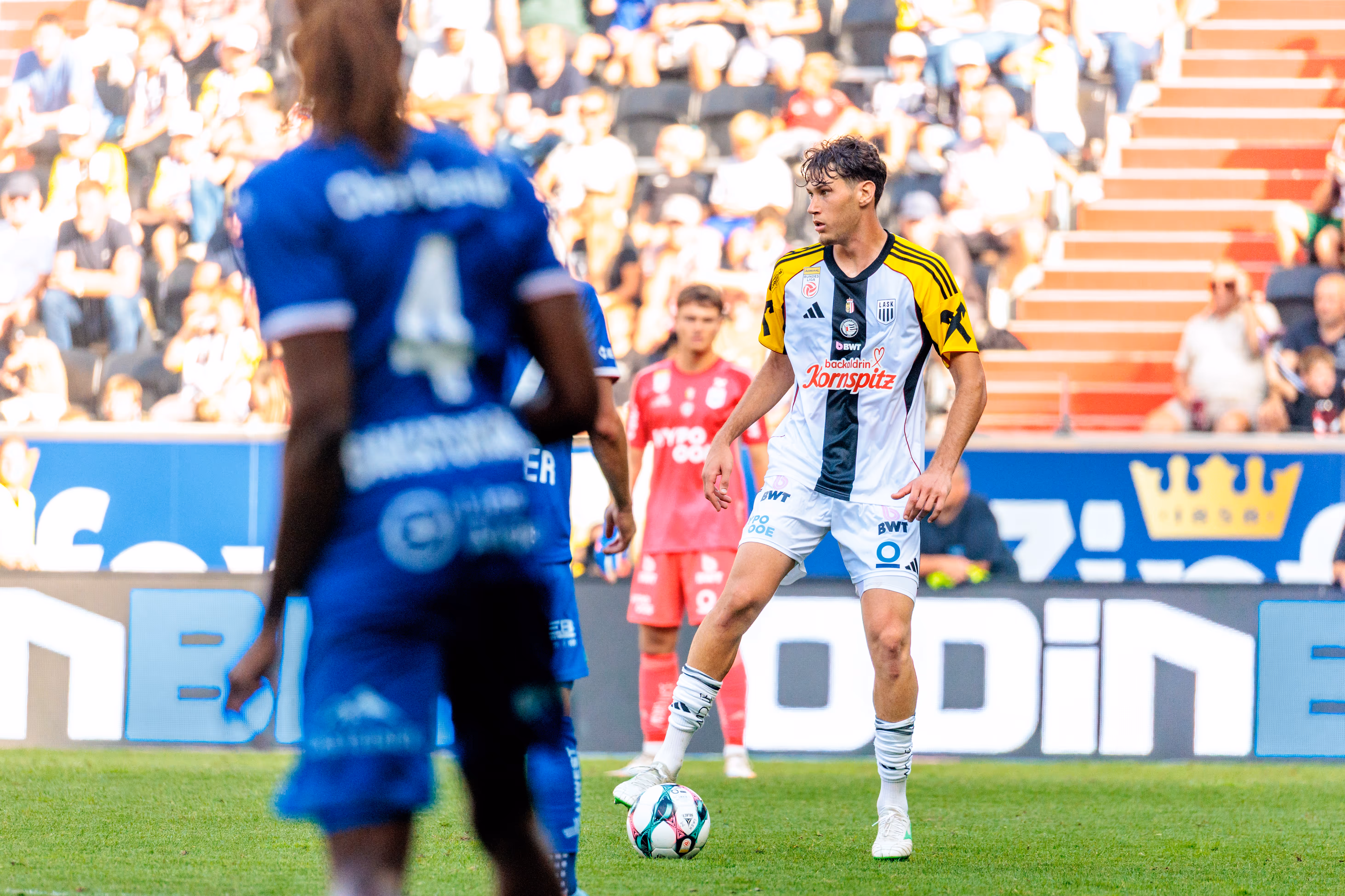 Ein Fußballspieler des LASK in weiß-schwarzem Trikot kontrolliert den Ball am Fuß, während ein Gegenspieler von Blau-Weiß Linz im unscharfen Vordergrund steht