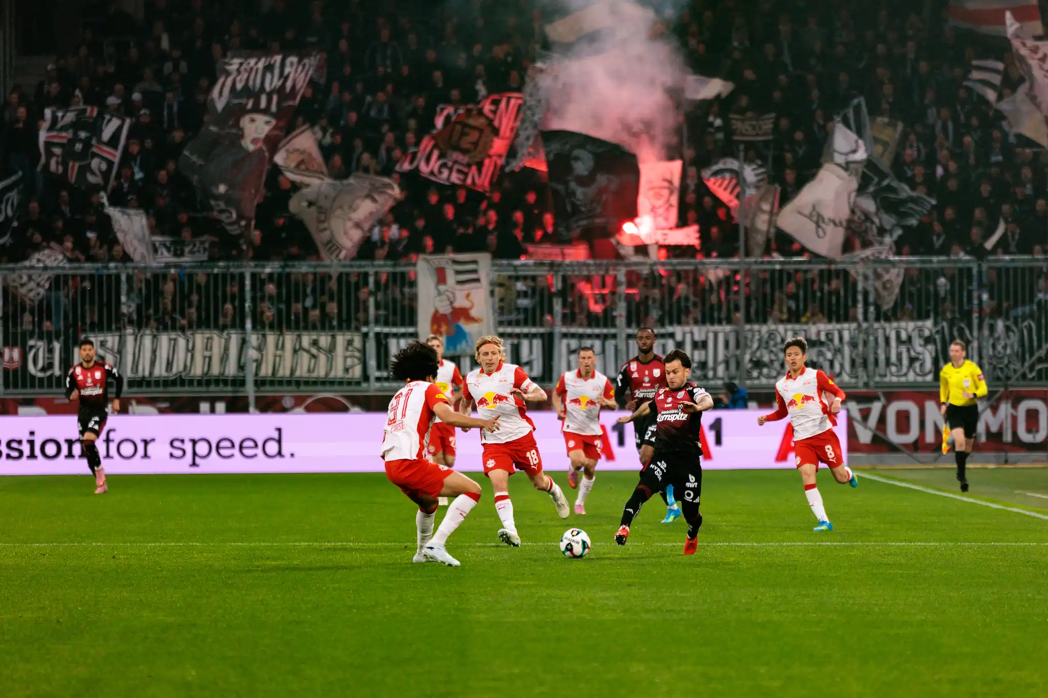 Ein Spieler des LASK führt den Ball im Dribbling gegen mehrere Verteidiger von Red Bull Salzburg vor einer mit Pyrotechnik und Fanbannern gefüllten Fankurve
