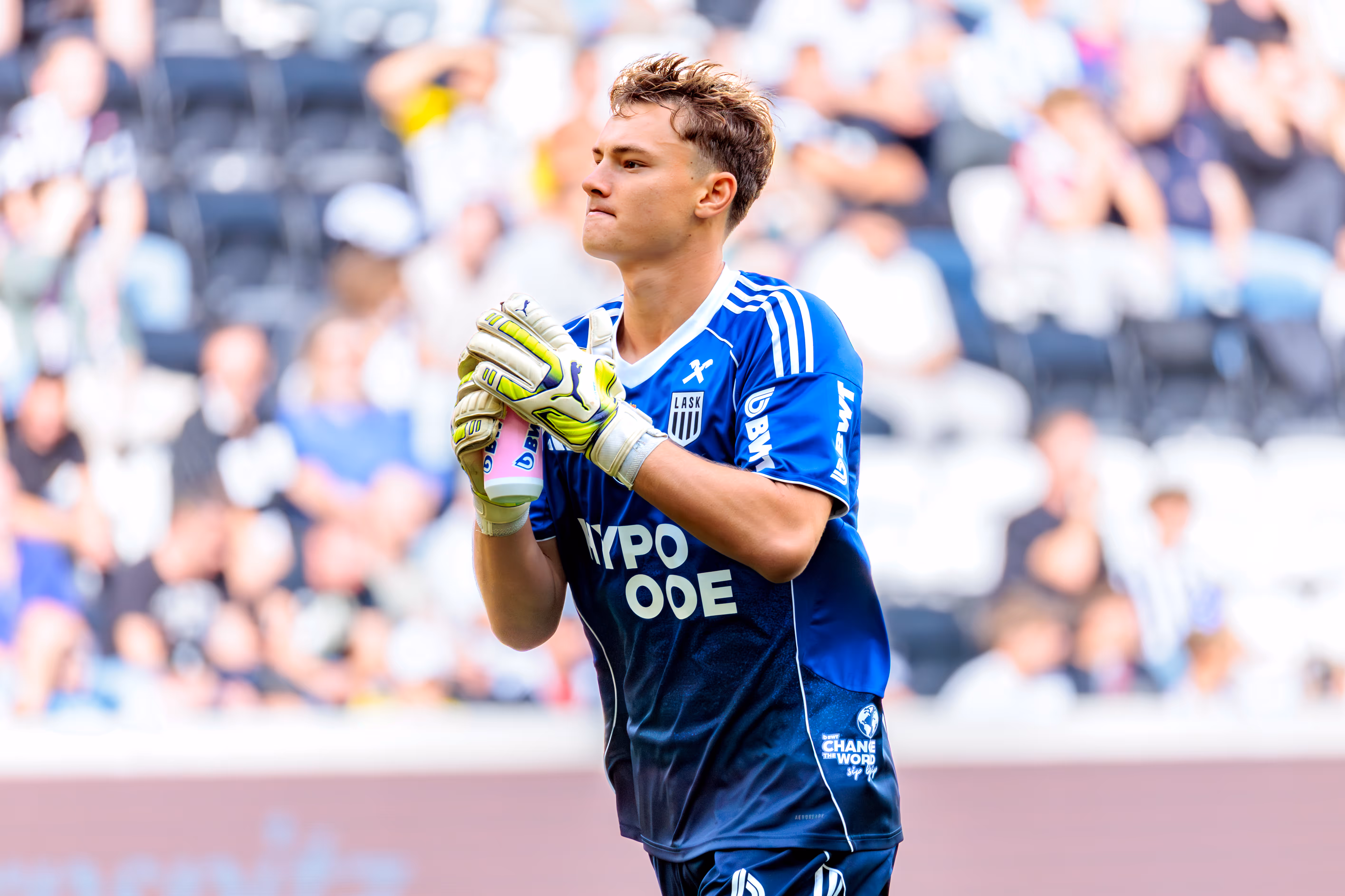 Lukas Jungwirth im blauen Heim-Torwart-Trikot mit HYPO OOE als Sponsor hält eine pinke BWT Trinkflasche und blickt während eines Bundesligaspiels zu den Fans auf während er vor dem Anpfiff zum Tor läuft