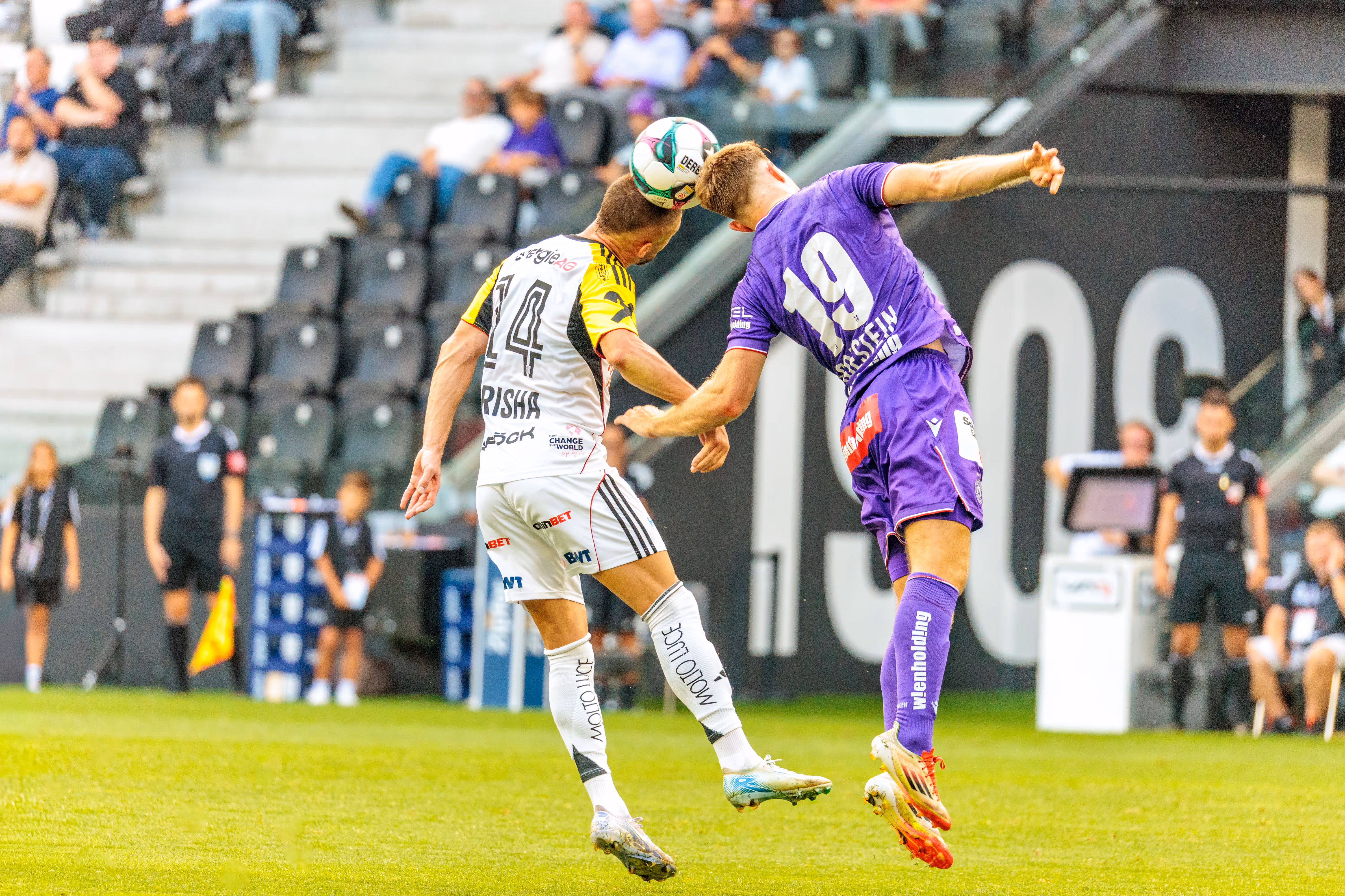Zwei Fußballspieler in schwarz-weißen und violetten Trikots springen für ein Kopfballduell in die Luft