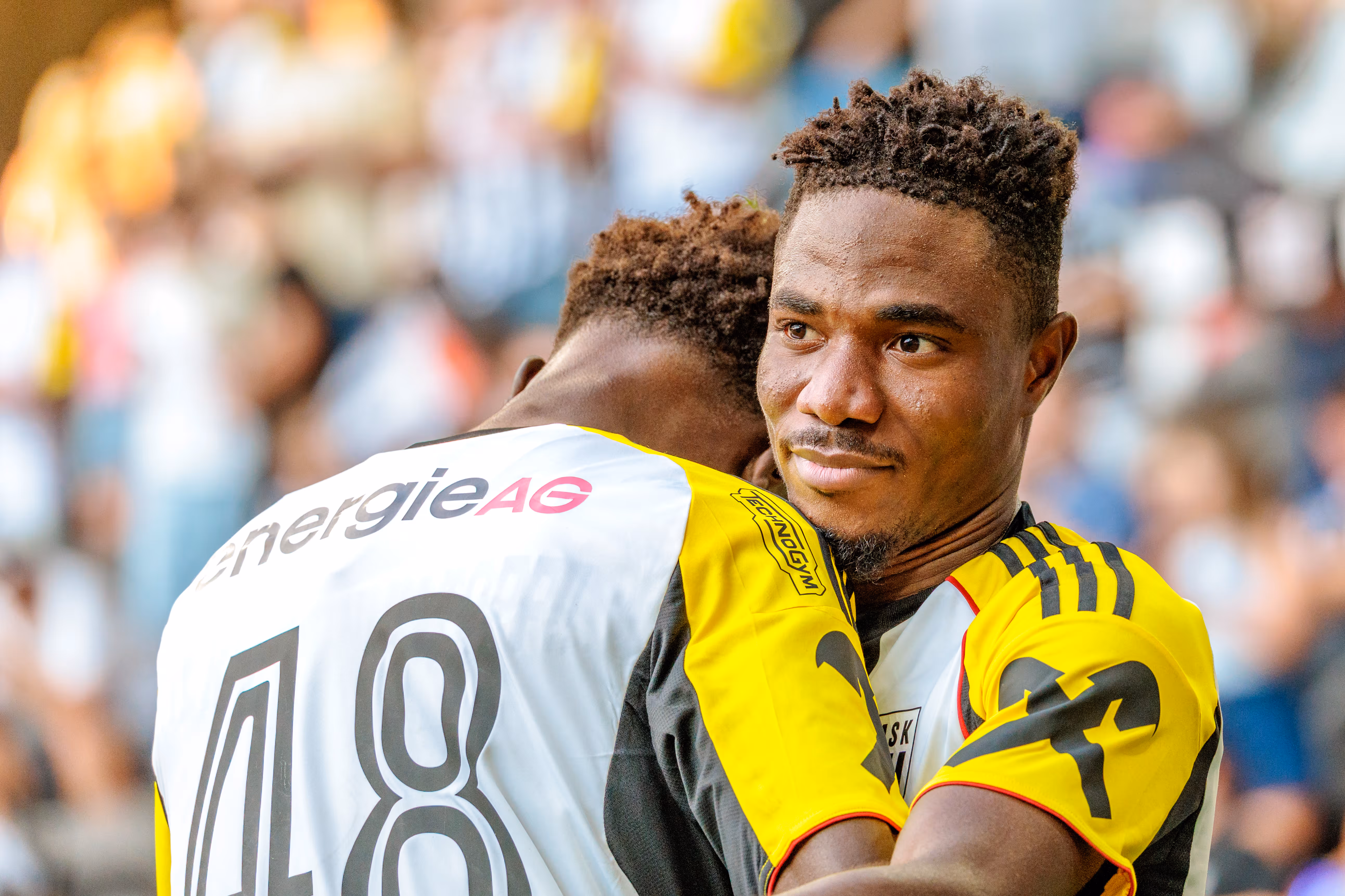 LASK-Fußballspieler Moses Usor im gelb-schwarzen Trikot blickt lächelnd in die Kamera, während er einen Teamkollegen in weißem Trikot umarmt