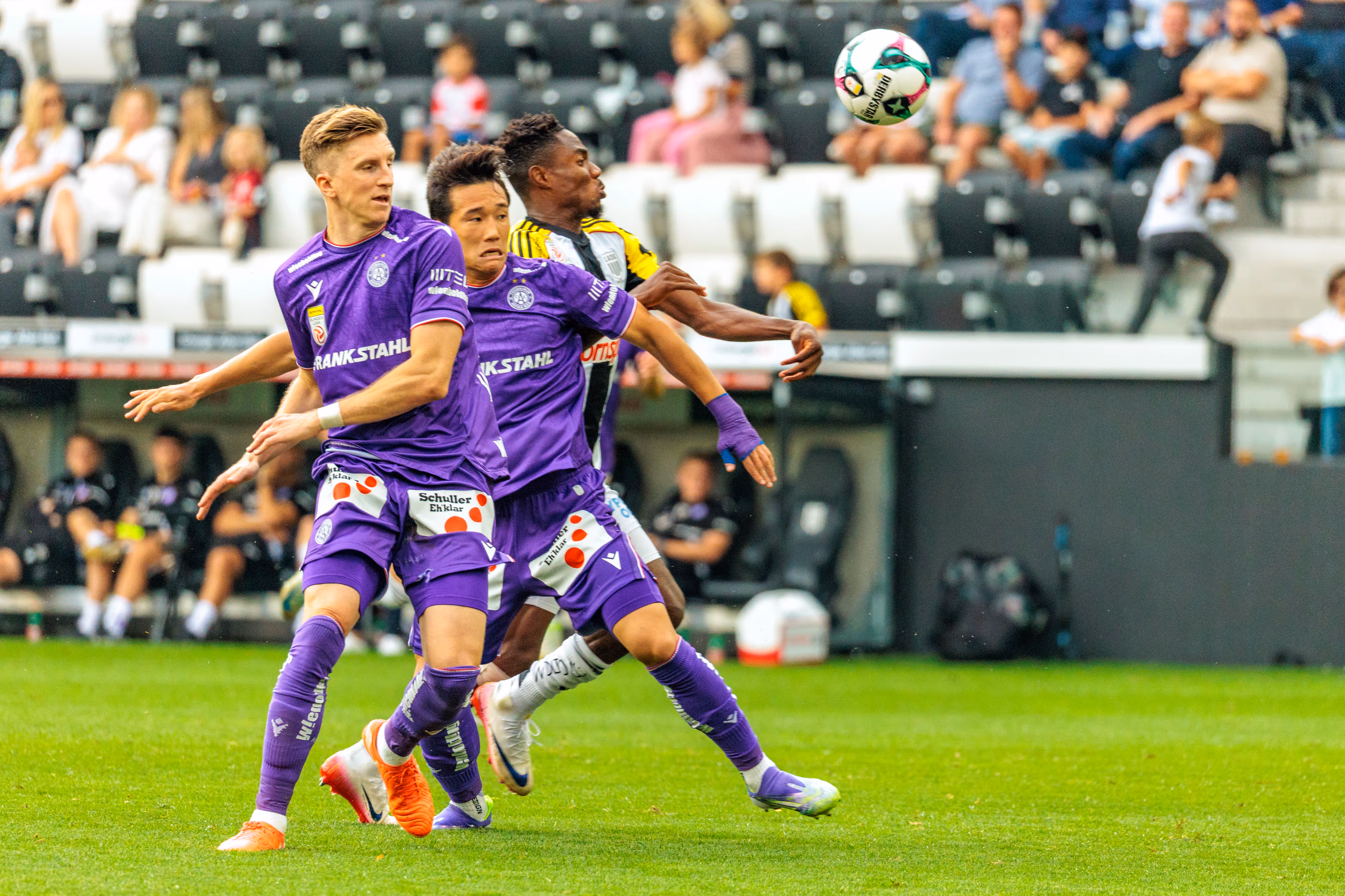Ein intensiver Zweikampf zwischen drei Fußballspielern, zwei in violetten und einem in einem schwarz-gelben Trikot, um den schwebenden Ball während eines Spiels der Bundesliga