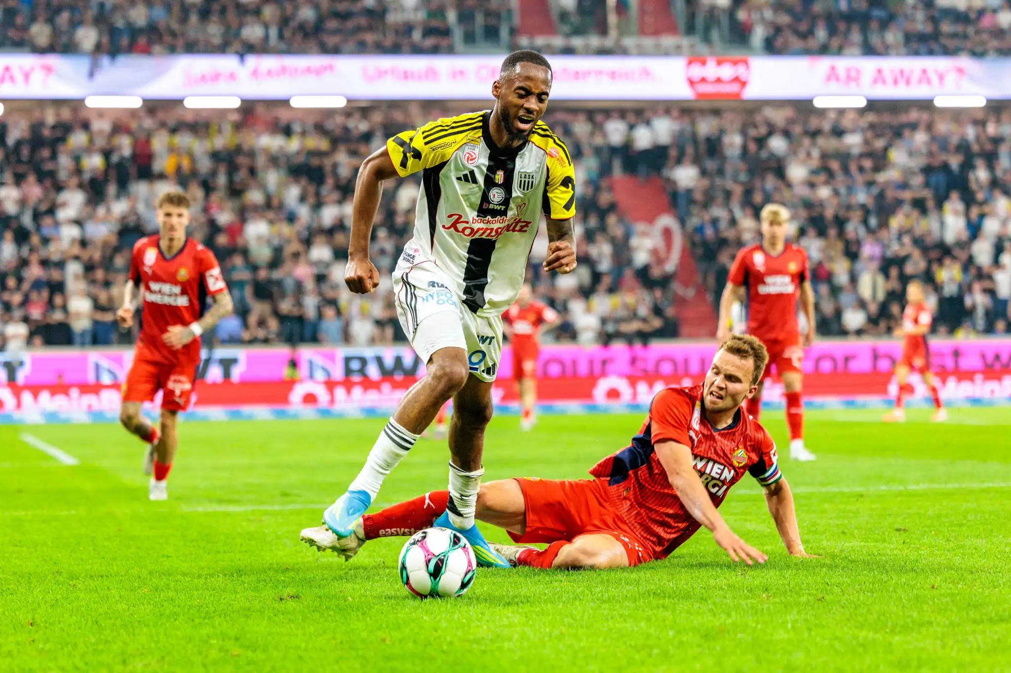 Ein Spieler des LASK führt den Ball an einem am Boden grätschenden Spieler des SK Rapid vorbei