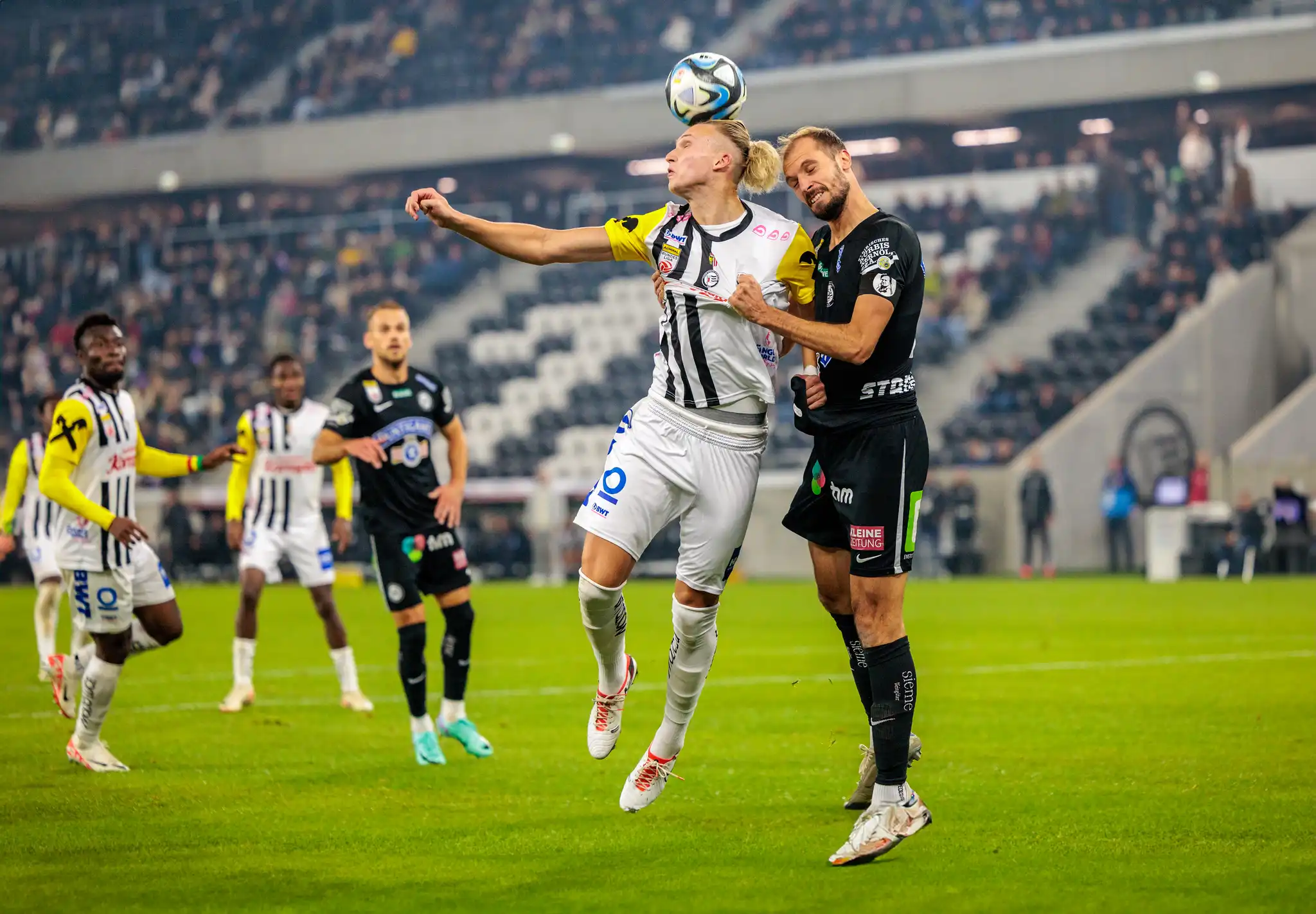 Ein LASK-Fußballspieler im schwarz-weiß gestreiften Trikot köpft den Ball im intensiven Zweikampf mit einem Gegenspieler im dunklen Trikot während eines Spiels in der Raiffeisen Arena 