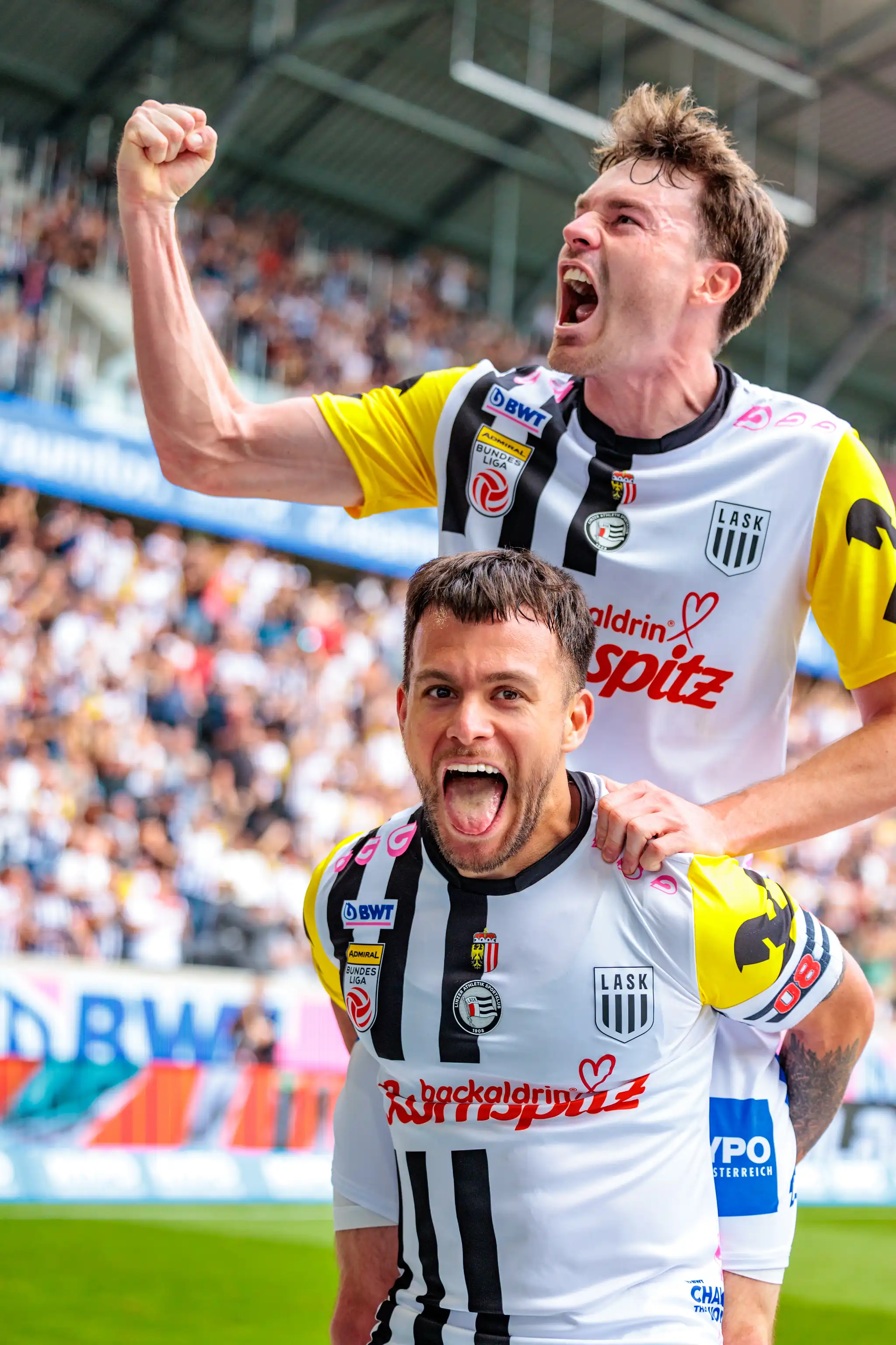 Zwei LASK-Spieler in ihren schwarz-weiß-gelben Trikots jubeln enthusiastisch in einem Fußballstadion 