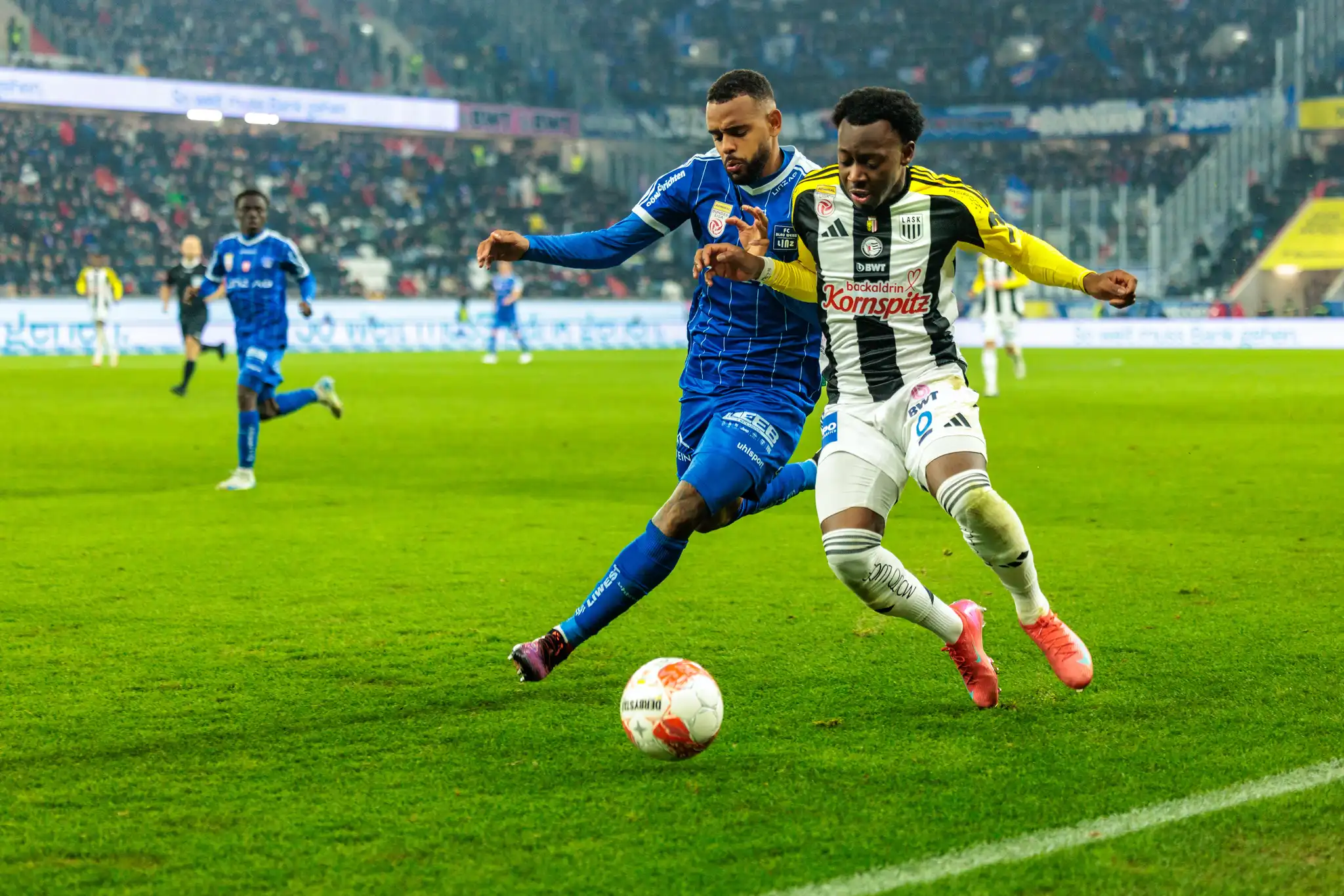 Zwei Fußballspieler, einer im blauen Trikot und einer vom LASK im gestreiften Trikot, sind im Zweikampf um den Ball auf dem Spielfeld der Raiffeisen Arena