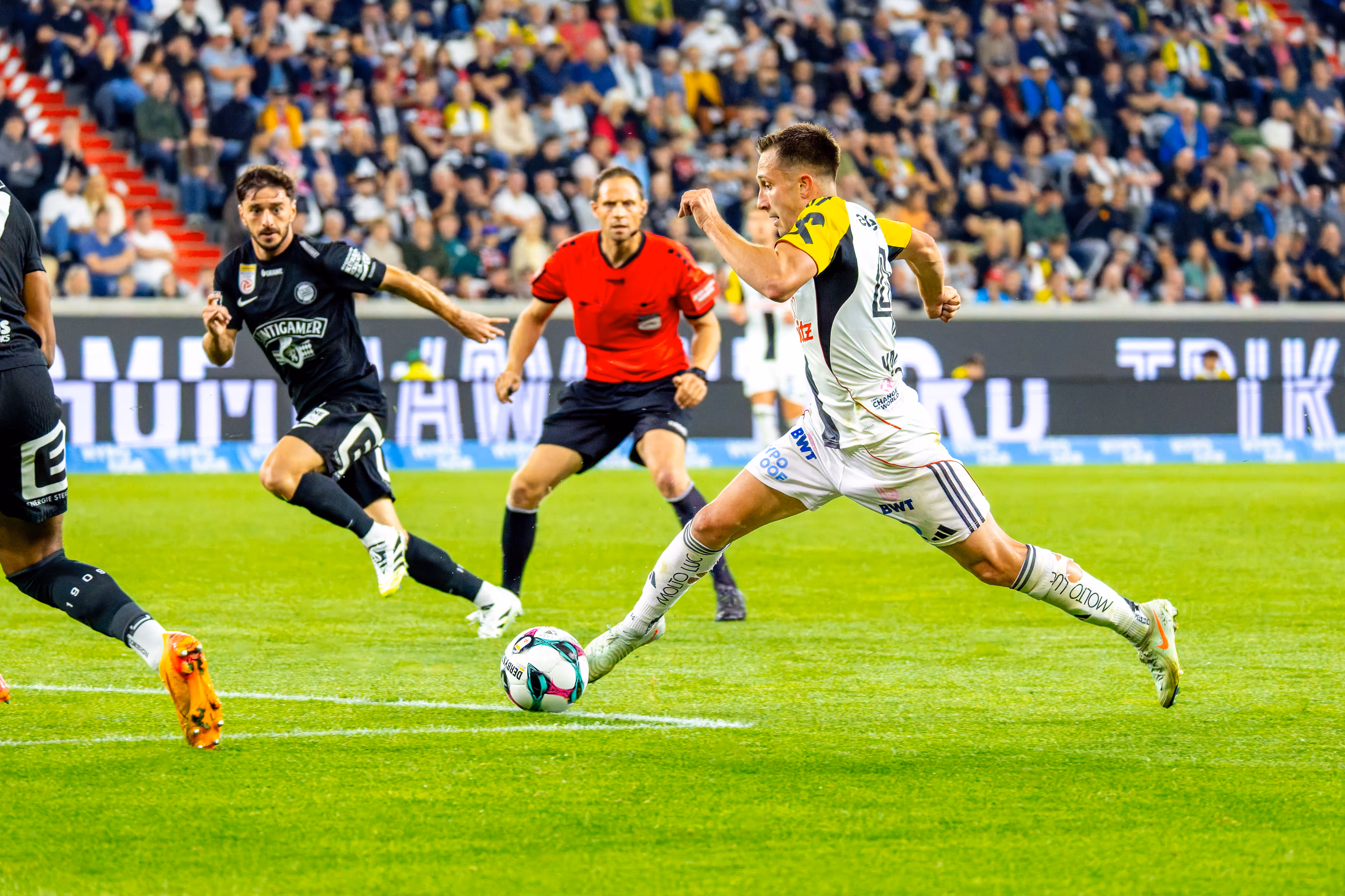 Dynamisches Bild eines Fußballspielers des LASK, der gerade einen Ball tritt, umgeben von einem Schiedsrichter, einem Gegenspieler und Zuschauern in der Raiffeisen Arena