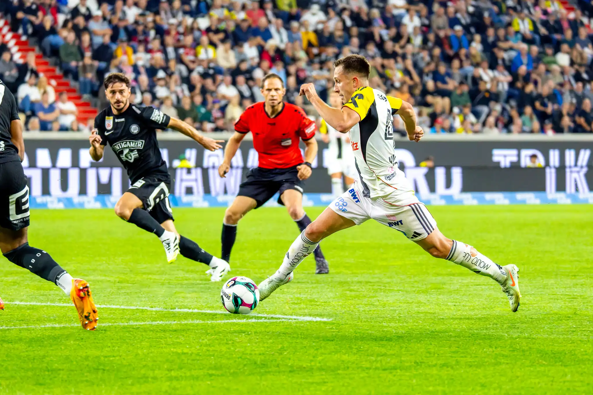 Dynamisches Bild eines Fußballspielers des LASK, der gerade einen Ball tritt, umgeben von einem Schiedsrichter, einem Gegenspieler und Zuschauern in der Raiffeisen Arena