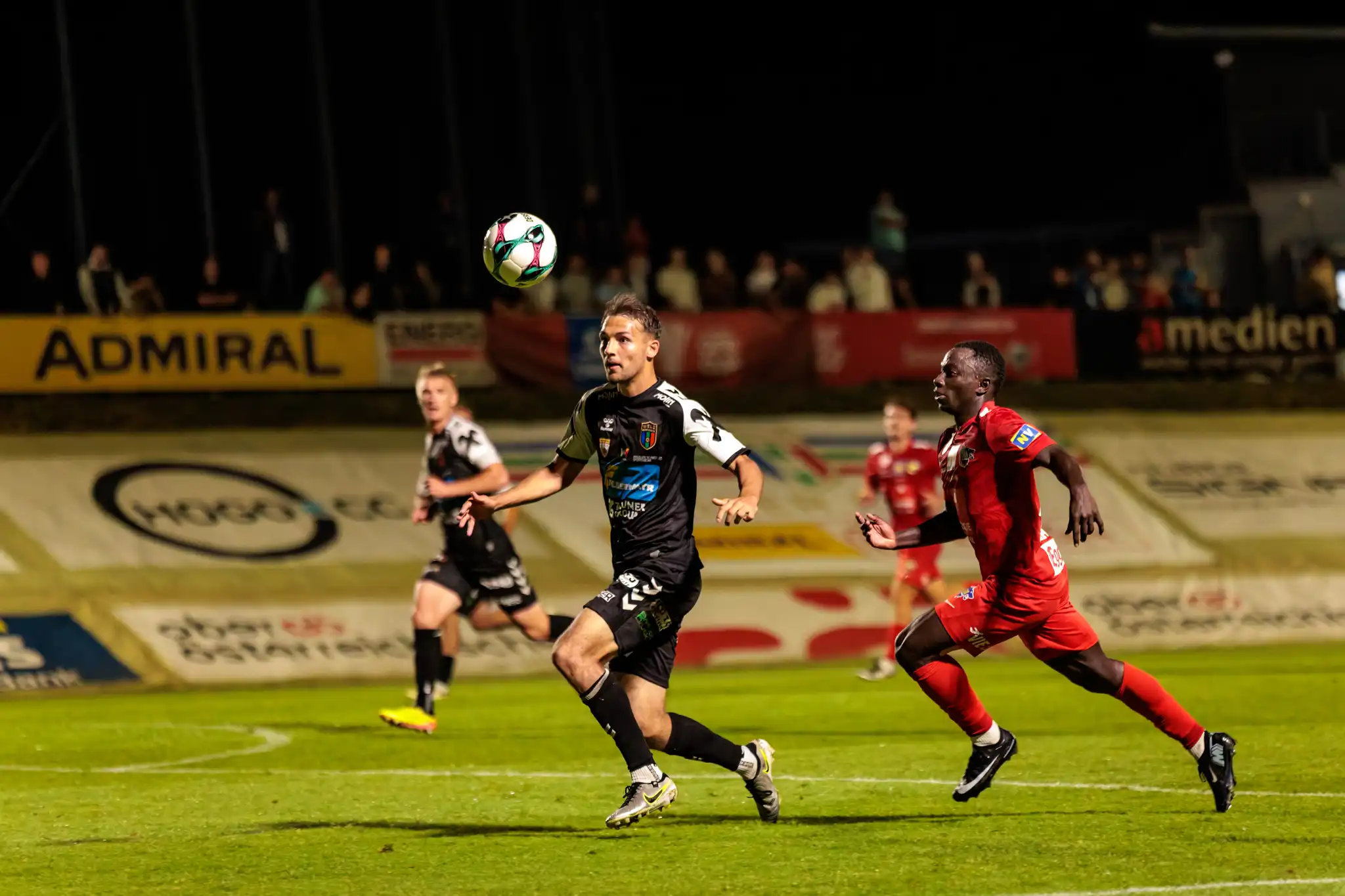 Ein Spieler in schwarzem Trikot und ein Spieler in rotem Trikot liefern sich ein intensives Laufduell um den Ball bei einem Flutlicht-Pokalspiel