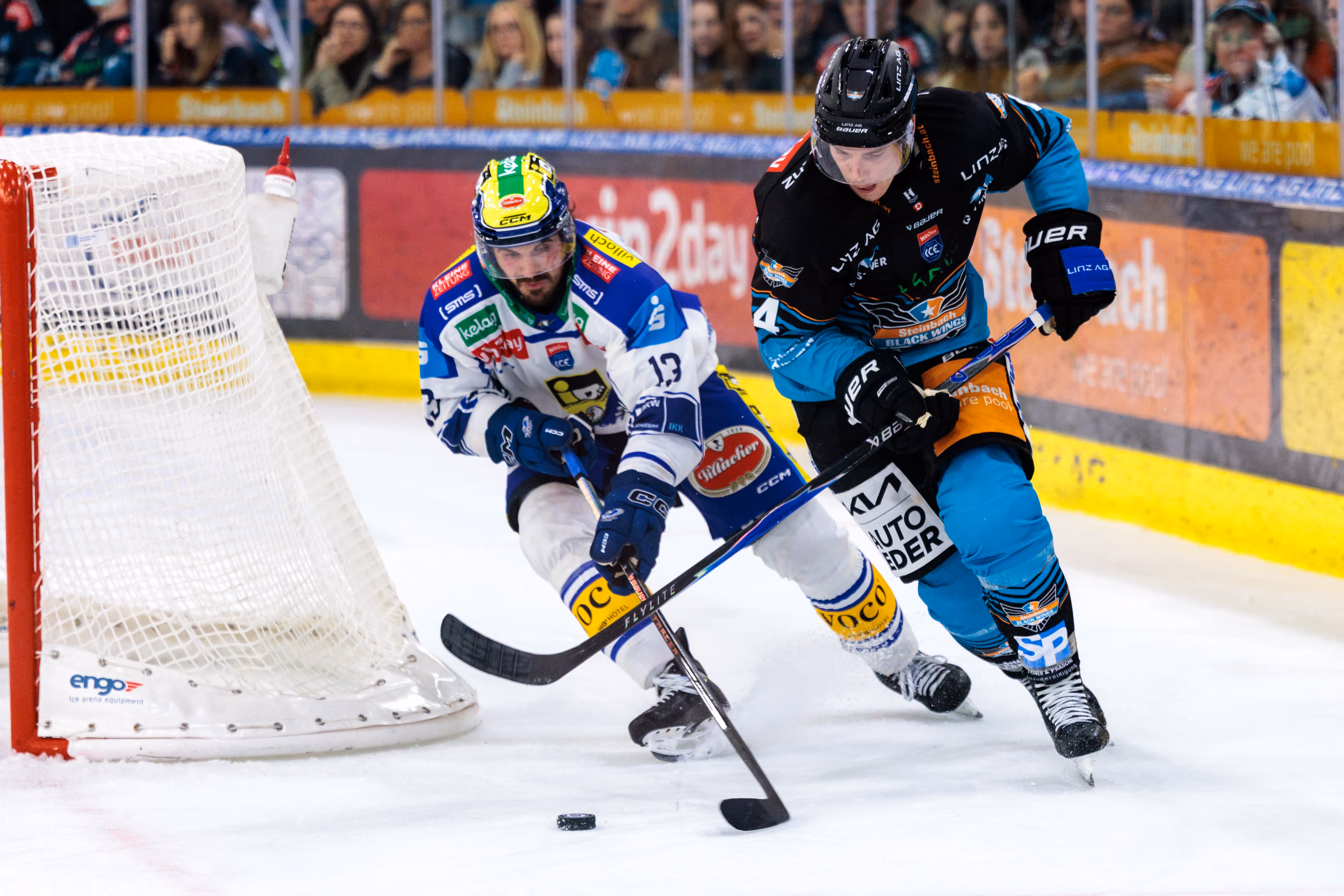 Eishockey-Spieler Adam Helewka (VSV, Nr. 13) und Ryan MacKinnon (Black Wings Linz, Nr. 4) kämpfen auf dem Eis vor dem Tor um den Puck
