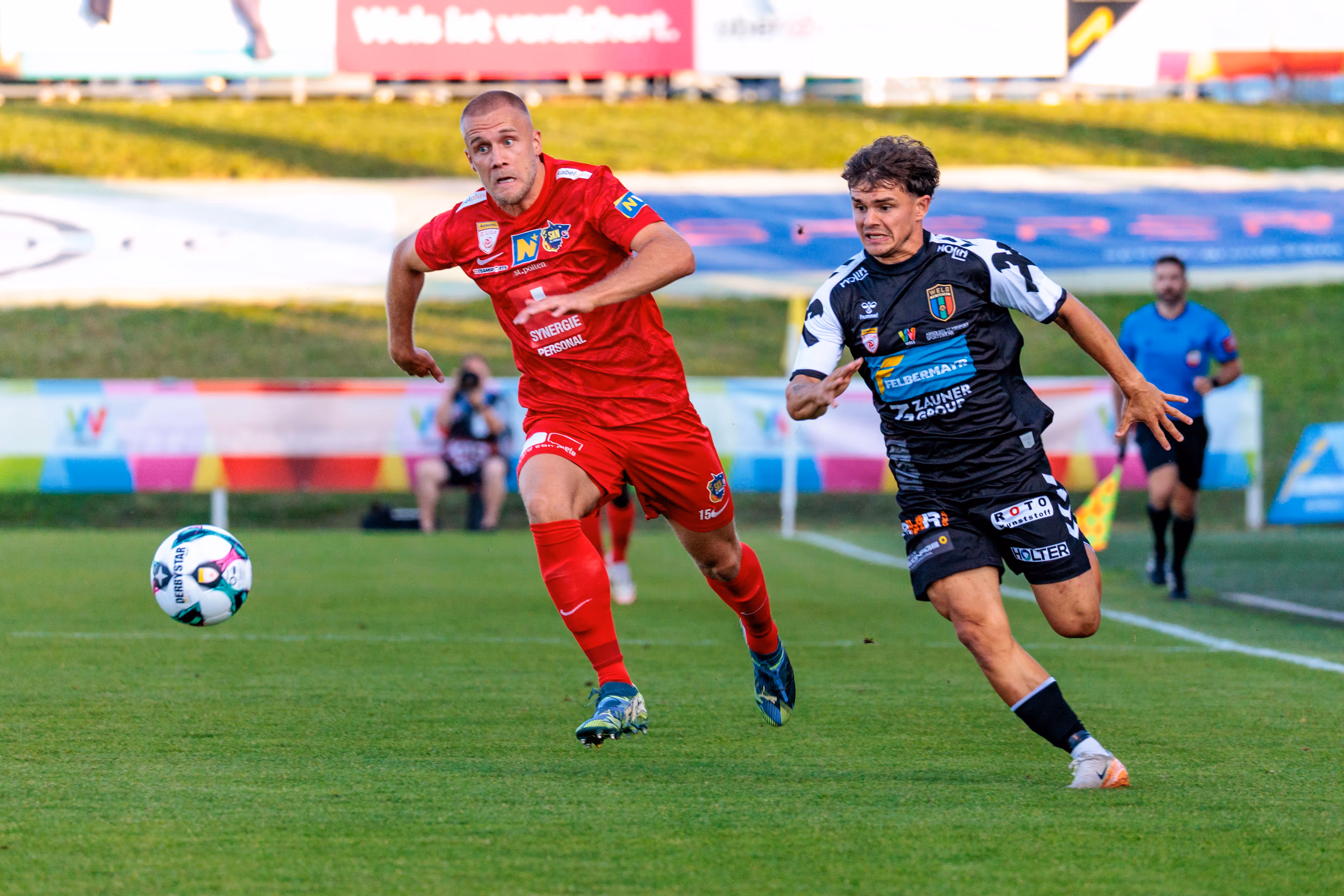 Im ÖFB Cup kämpfen ein Spieler des SKN St. Pölten in rotem Trikot und ein Kontrahent von Herta Wels in schwarz-weißem Dress energisch um den Ball