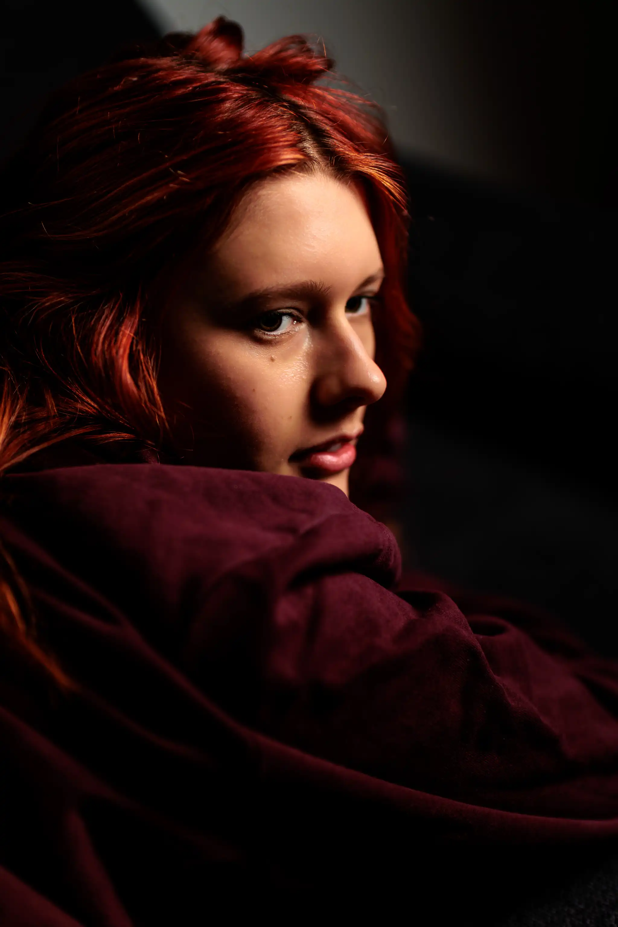 Nahaufnahme eines ausdrucksstarken Beauty-Portraits der rothaarigen Sarah in bordeauxrotem Hoodie, die mit intensivem Blick über die Schulter im stimmungsvollen Seitenlicht posiert