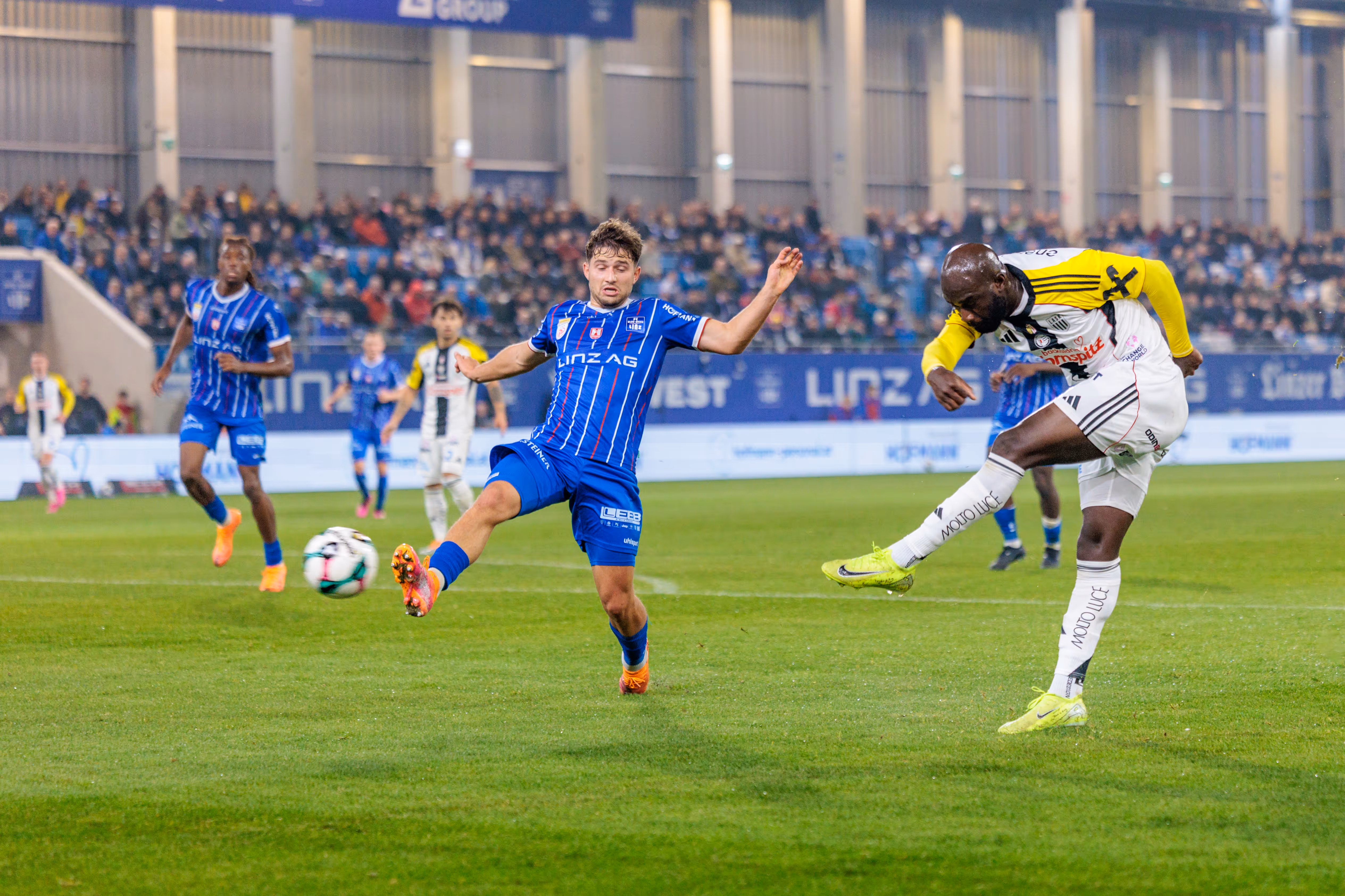 LASK-Spieler Moses Adeniran schießt den Ball mit energischem Einsatz auf das Tor, während ein Spieler von Blau-Weiß Linz daneben steht