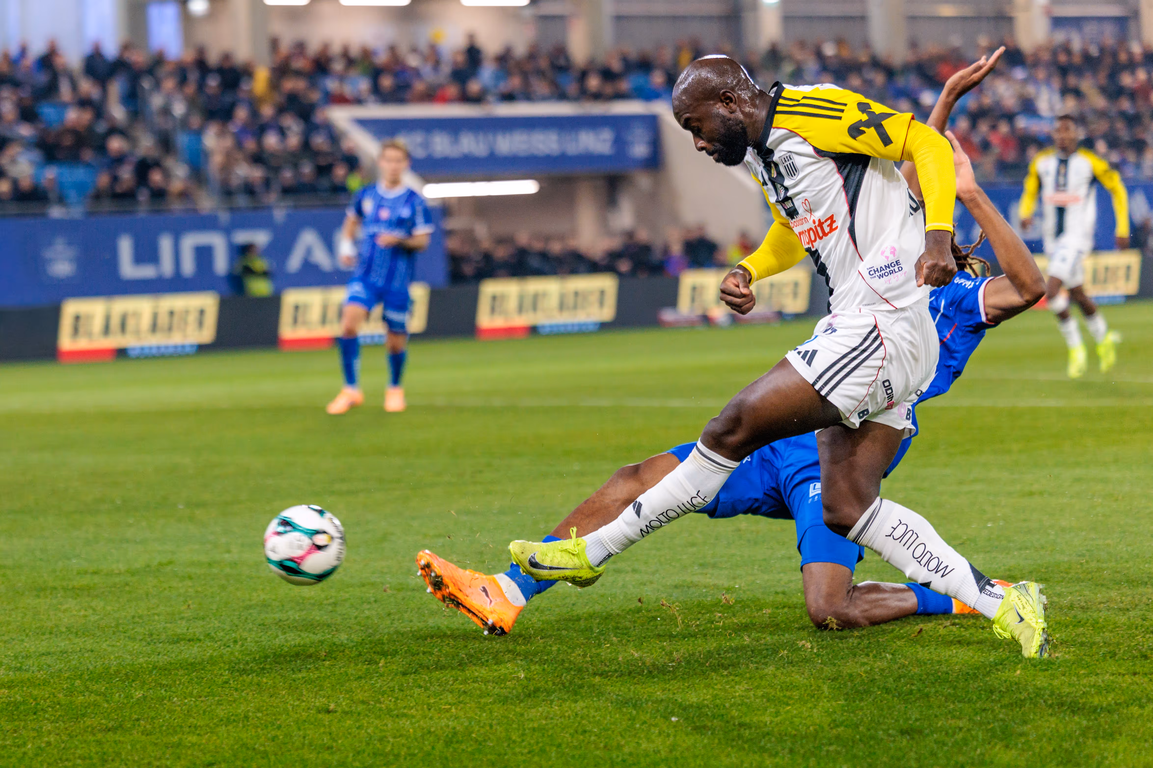 Moses Adeniran im gelb-weißen LASK-Trikot gibt einen Torschuss ab, während ein Spieler von Blau-Weiß Linz in blauem Trikot am Boden liegend blockt