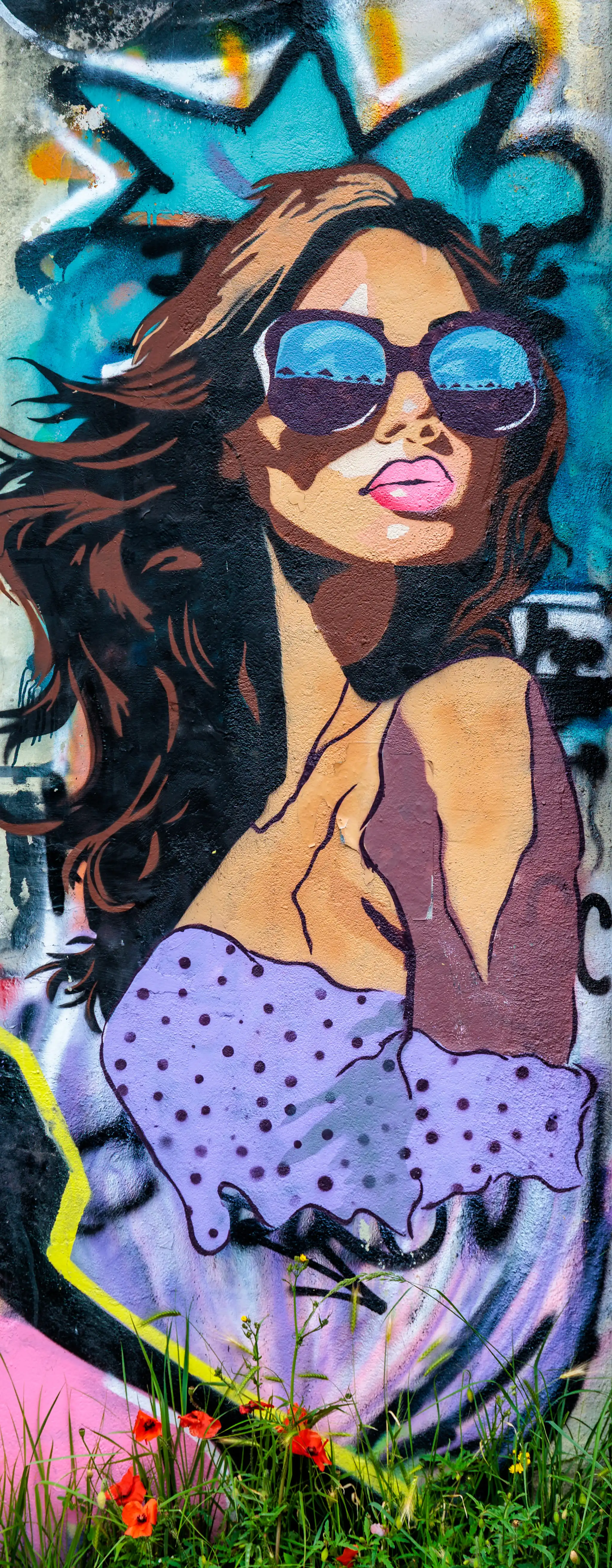 Ein farbenfrohes Graffiti-Wandbild einer Frau mit Sonnenbrille und wehendem Haar ziert eine Mauer, im Vordergrund wachsen grüne Pflanzen und rote Mohnblumen