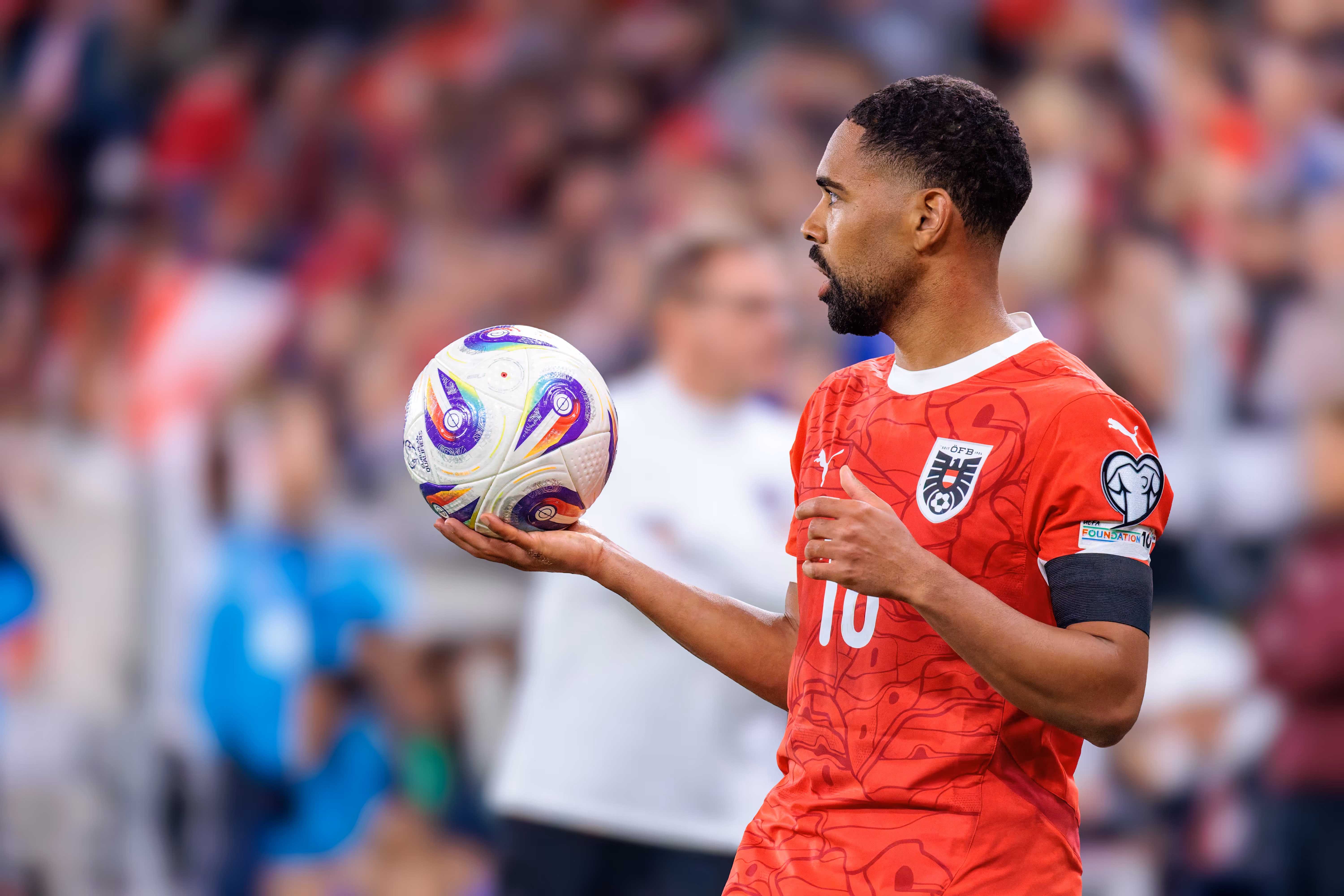 Der österreichische Nationalspieler Phillipp Mwene im roten Trikot mit der Rückennummer 10 hält einen offiziellen UEFA European Qualifiers Fußball in der Hand und blickt zu den Mitspielern