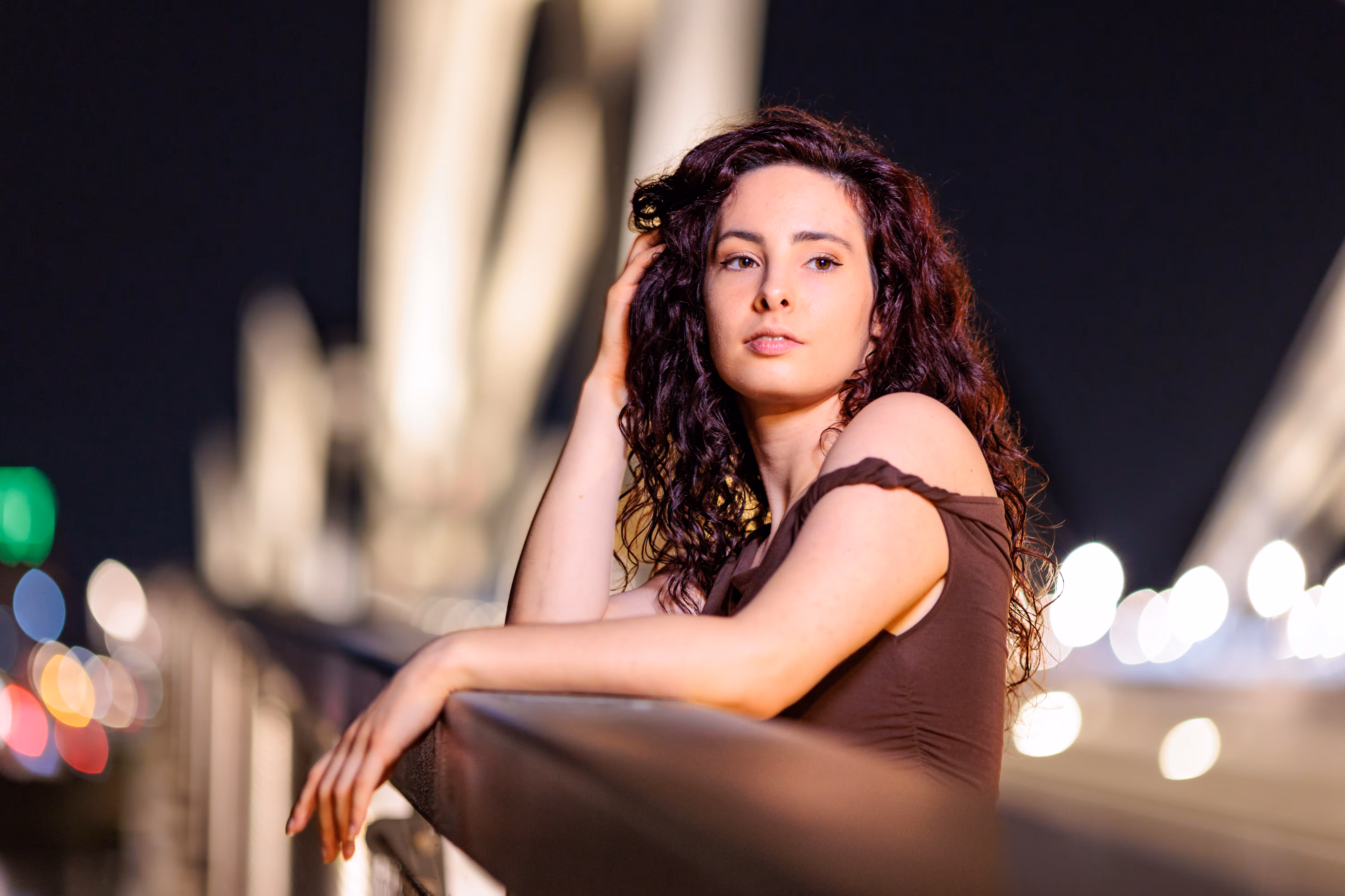 Porträt von Nicole mit dunklem, lockigem Haar, die bei Nacht auf der Linzer Eisenbahnbrücke anlehnt, im Hintergrund verschwimmen stimmungsvolle Stadtlichter