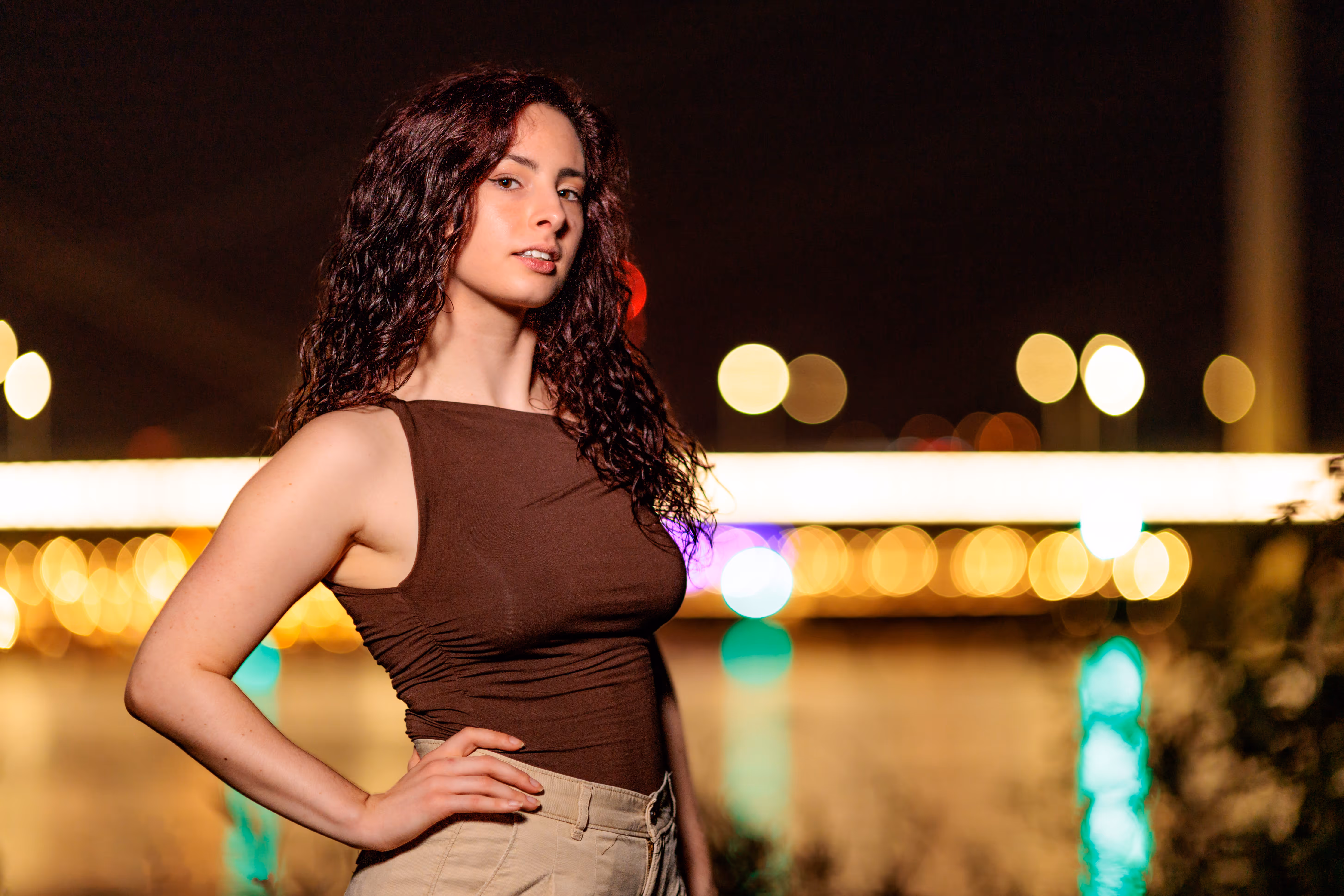Ein Portrait von Nicole mit lockigem Haar in einem braunen Oberteil, selbstbewusst stehend vor dem nächtlichen Linz mit seinen funkelnden Bokeh-Lichtern