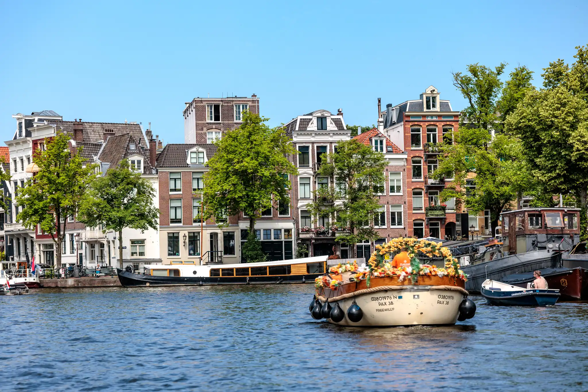 Ein malerisches Blumenboot mit bunten orangefarbenen Blumen fährt auf einem Kanal in Amsterdam, umgeben von traditionellen Grachtenhäusern und Bäumen unter blauem Himmel