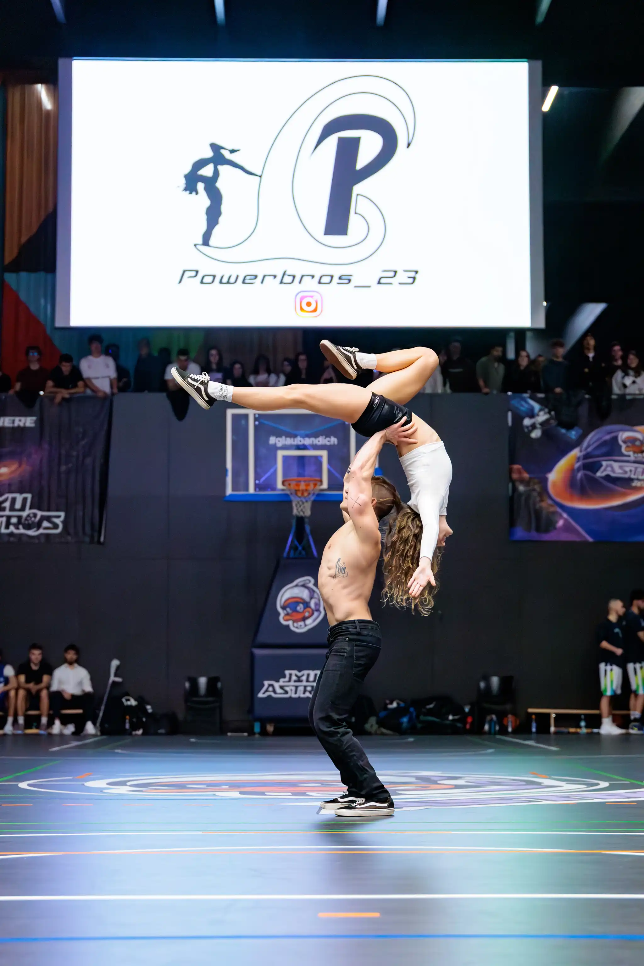 Eine Turnvorführung präsentiert einen Mann, der eine Frau in einer eleganten Hebefigur in einer Sporthalle mit großem "Powerbros" Bildschirm trägt