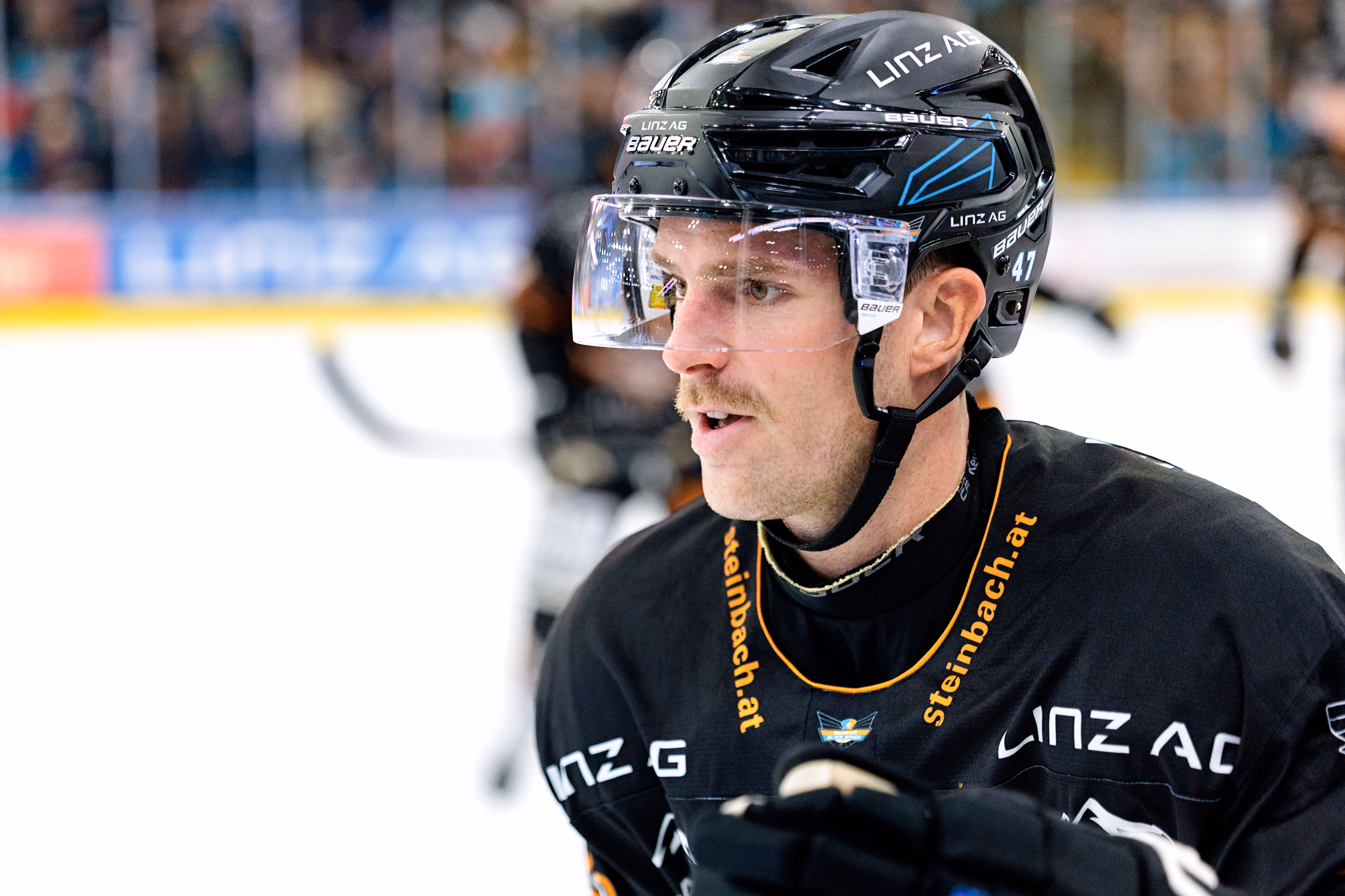 Eine Porträtaufnahme des Eishockeyspielers Logan Roe von den Black Wings Linz, der während eines Spiels in seiner schwarzen Ausrüstung konzentriert zur Seite blickt