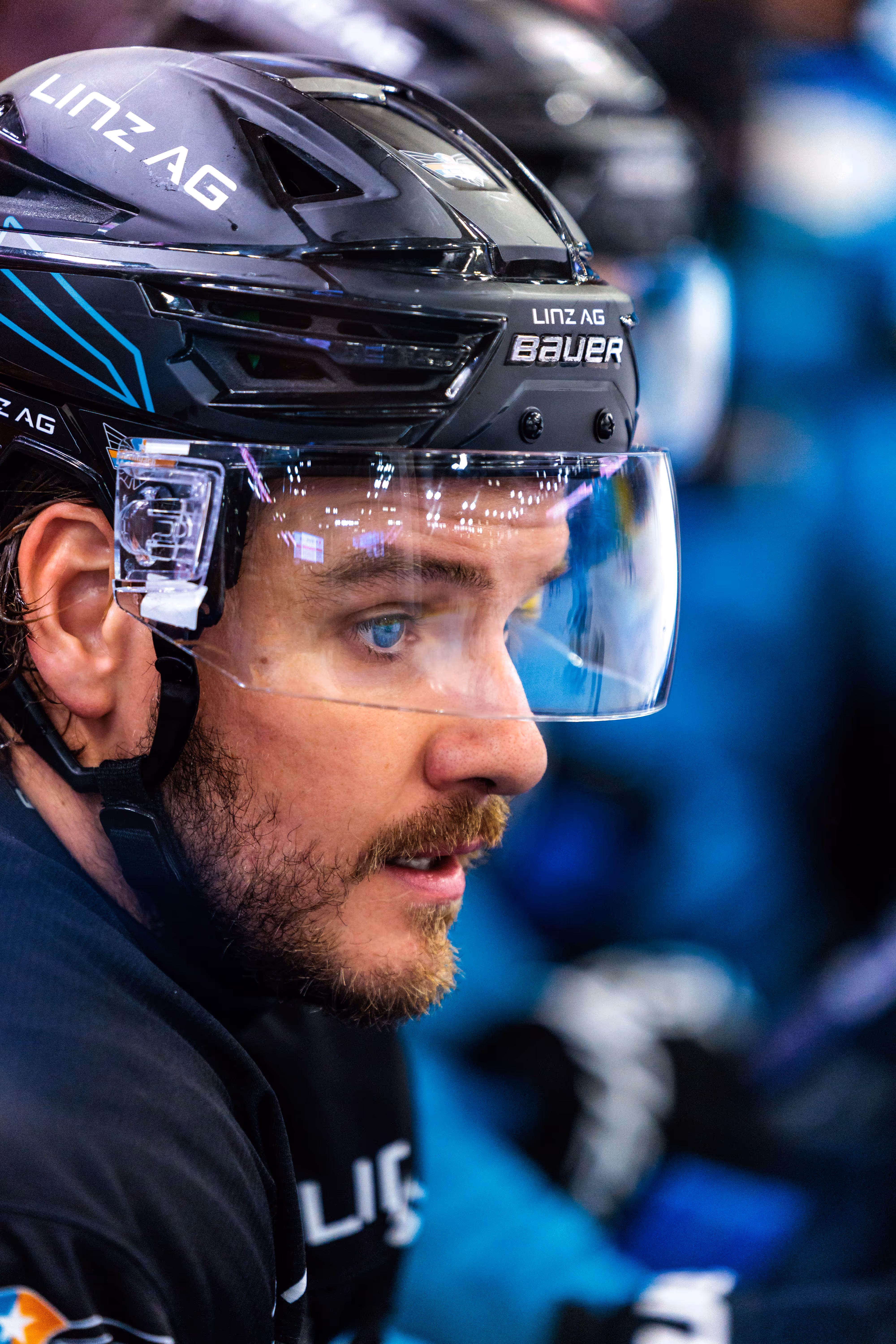 Der Eishockeyspieler Travis Barron blickt fokussiert zur Seite, während er seinen schwarzen Helm mit dem Linz AG-Logo und einem klaren Visier trägt