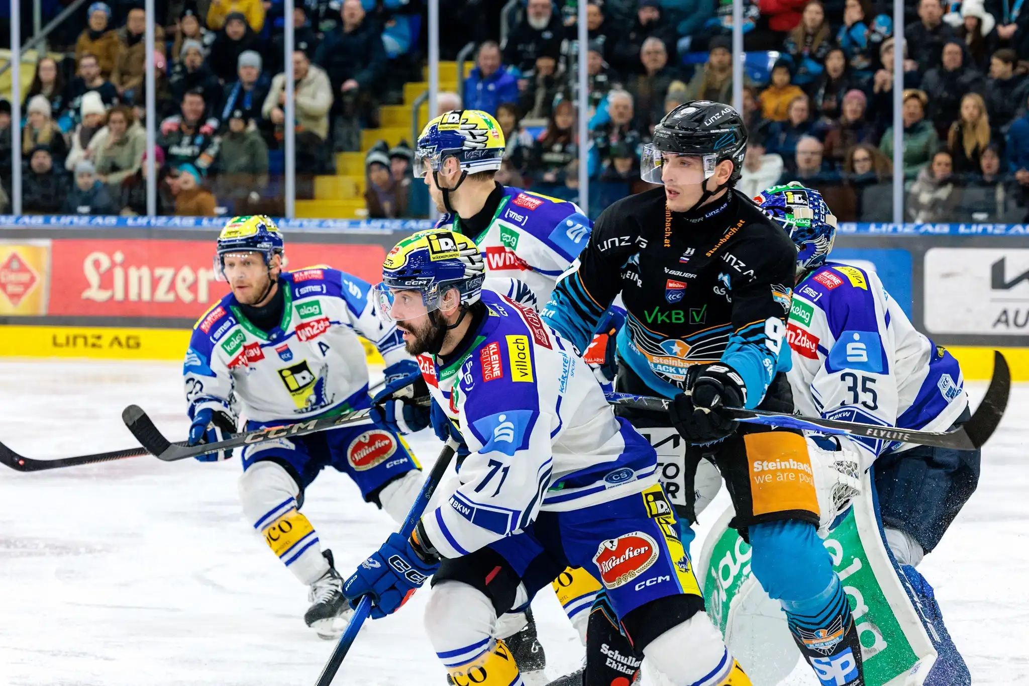 Eishockeyspieler der Black Wings Linz in schwarzen Trikots und des VSV in blauen Trikots kämpfen intensiv um den Puck auf dem Eis, während der Torhüter des VSV bereitsteht