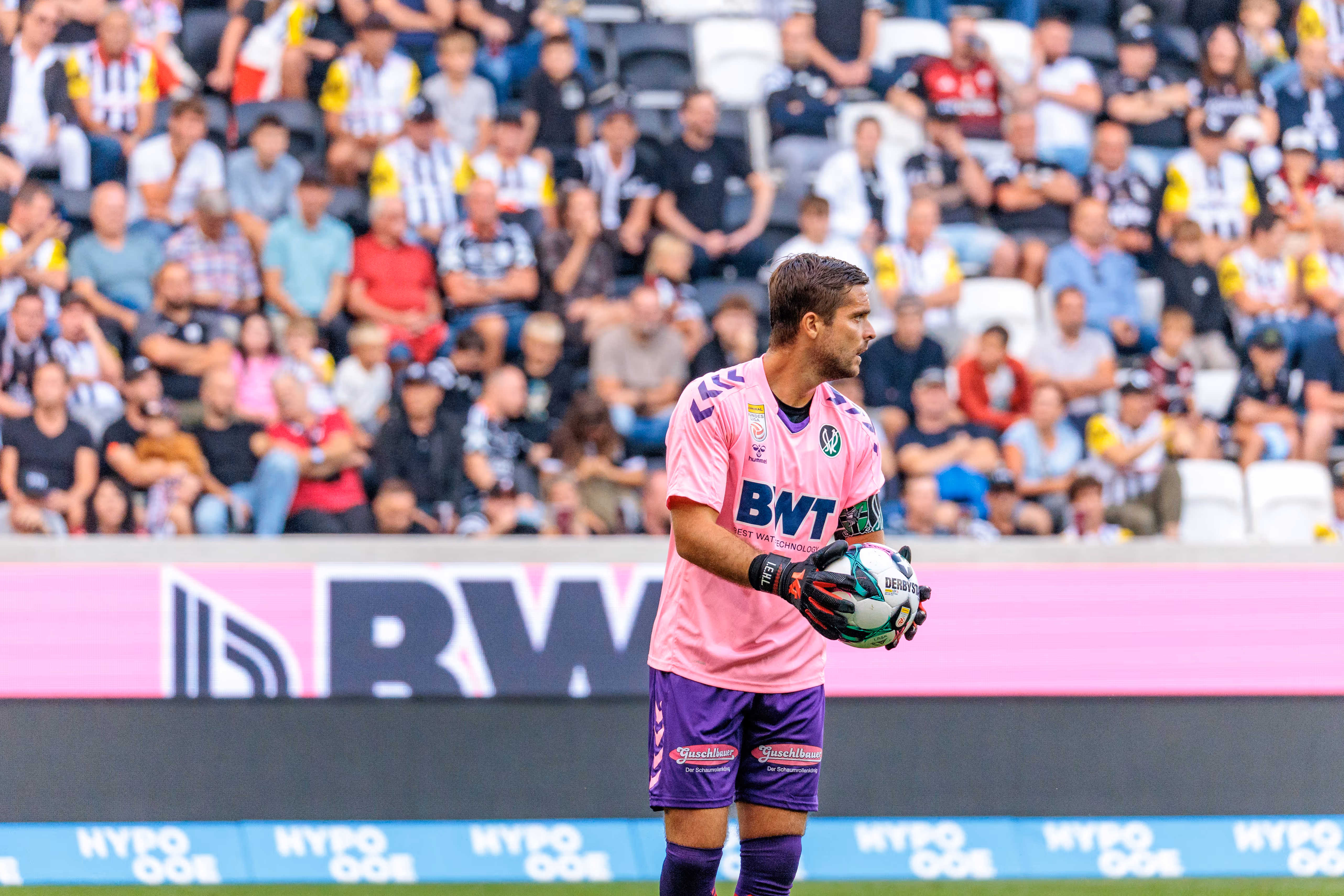 Amdreas Leitner im pinken BWT hält den Spielball während eines Derbyspiels gegen den LASK, mit unscharfen Fans und einer BWT Bande im Hintergrund