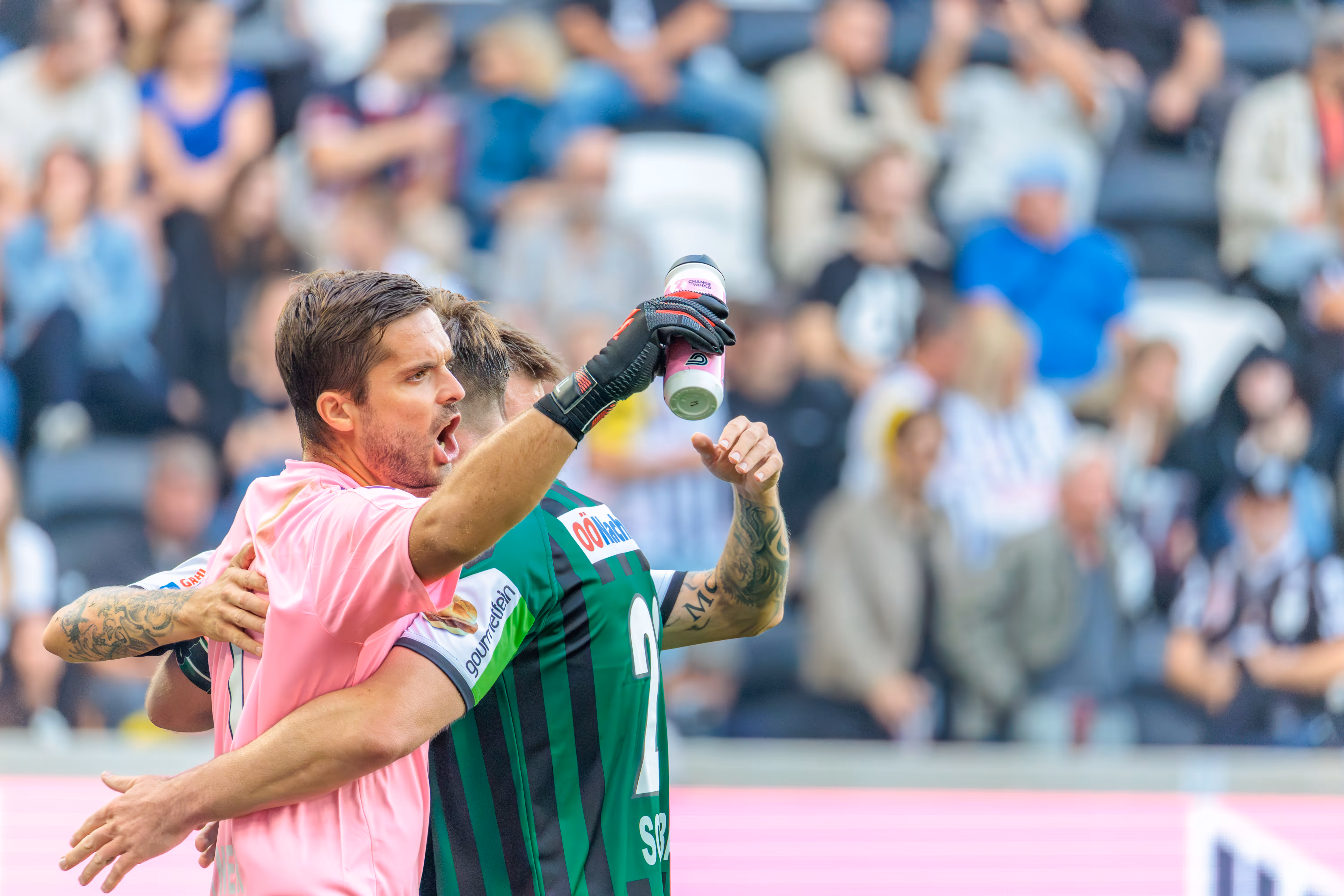 Der Torhüter des SV Ried in pinkem Trikot und Handschuhen jubelt mit einem Mitspieler in grün-schwarz gestreiftem Trikot über den Derby-Sieg gegen den LASK Linz