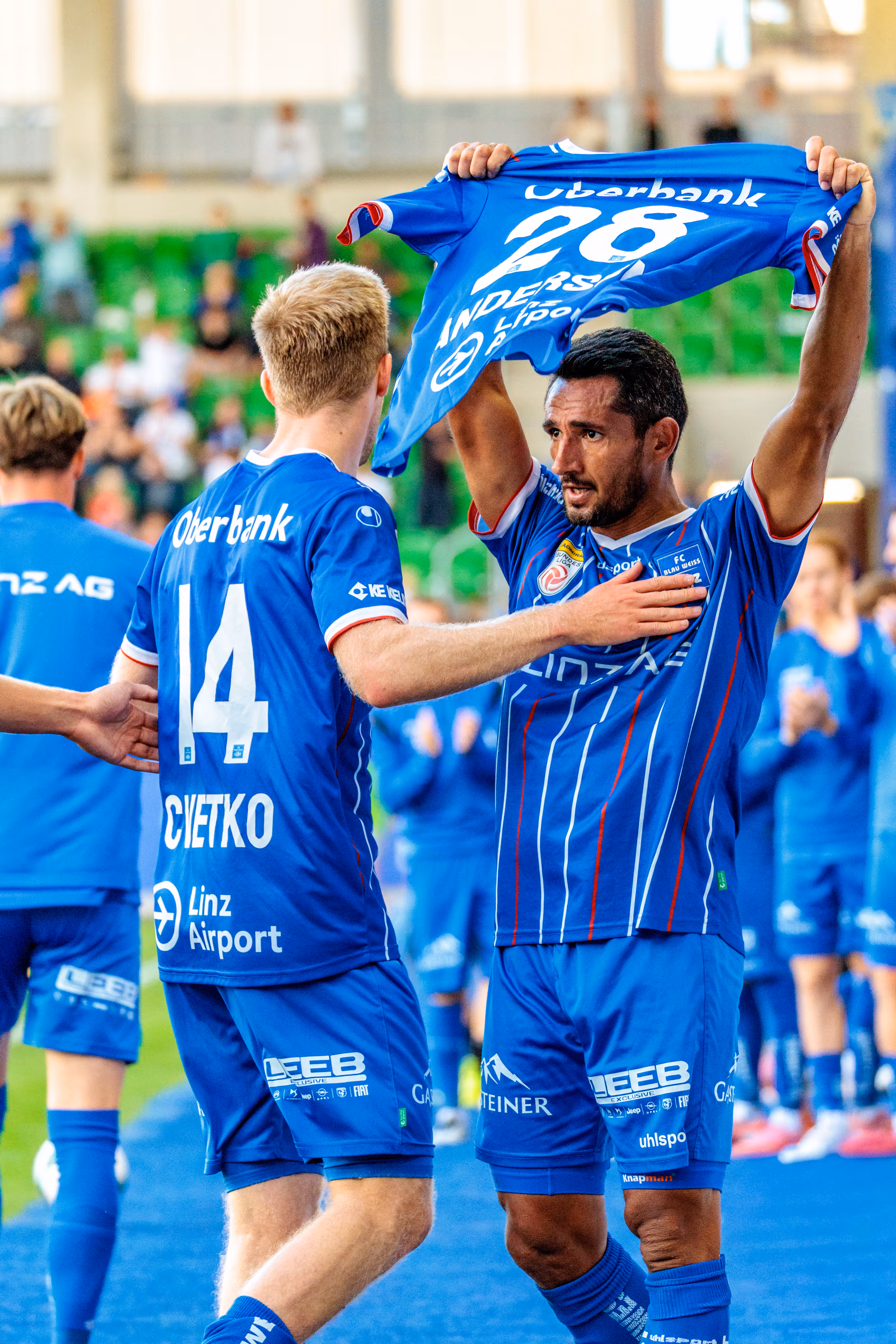 Der Fußballspieler Ronivaldo vom FC Blau-Weiß Linz hält nach seinem Tor emotional das Trikot des verletzten Anders Capek hoch, während sein Teamkollege Christopher Cvetko mit ihm feiert
